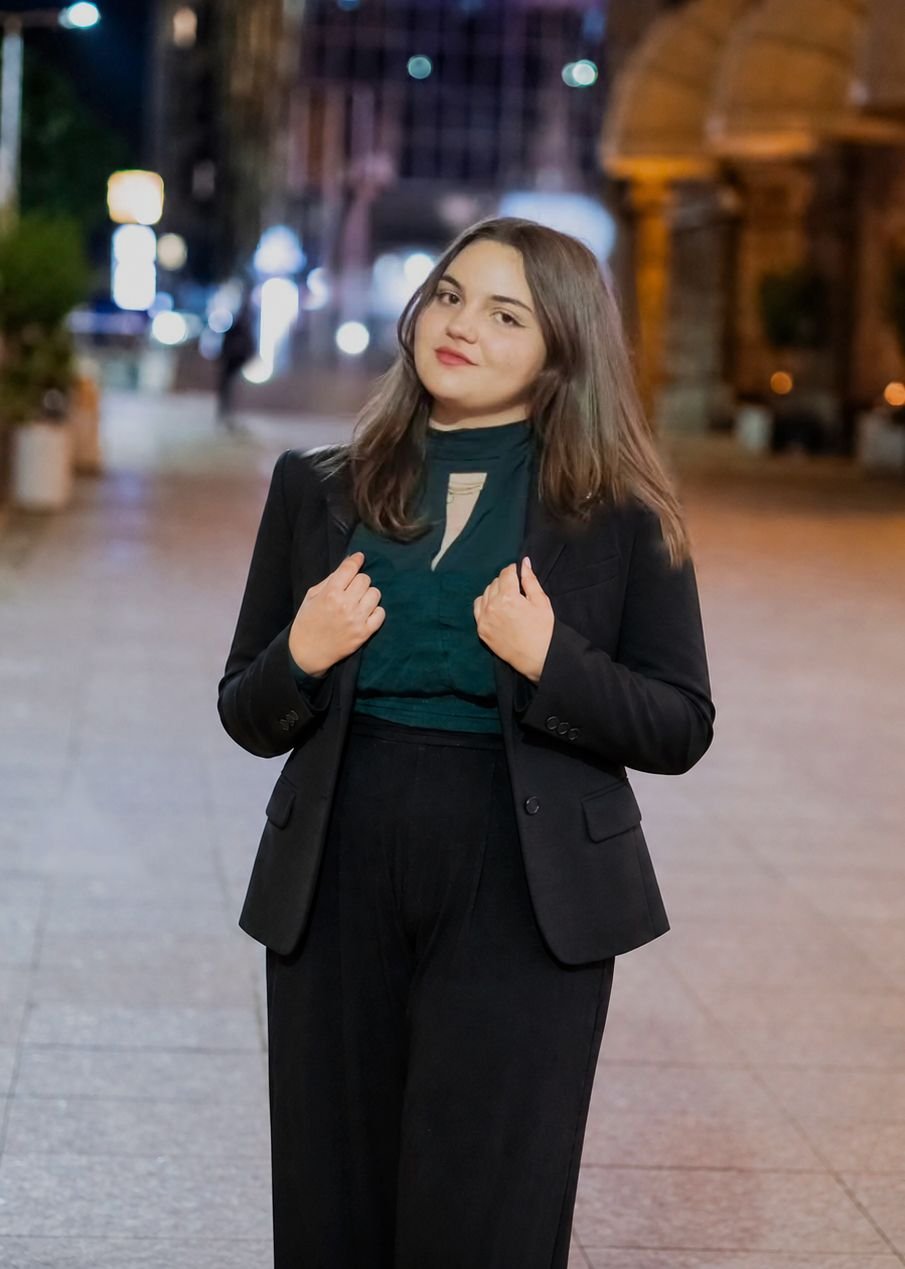 A young woman with shoulder-length brown hair standing on a city street at night, wearing a black blazer and pants, with a green top underneath, and posing confidently with a slight smile.