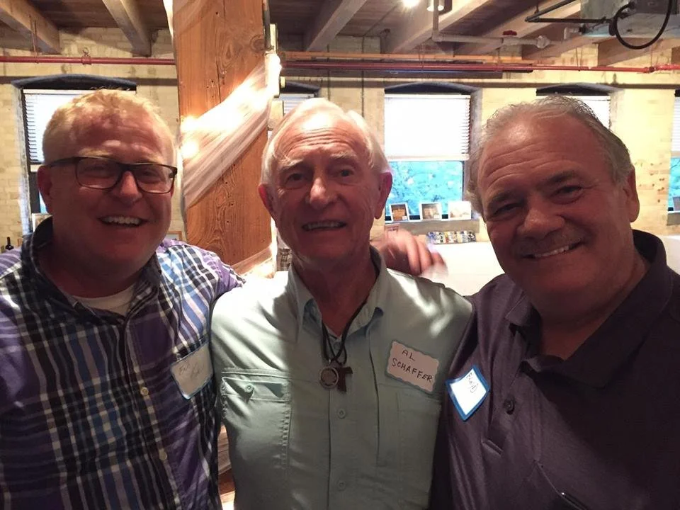 Three men standing close together smiling in an indoor setting with exposed brick walls and wooden beams.