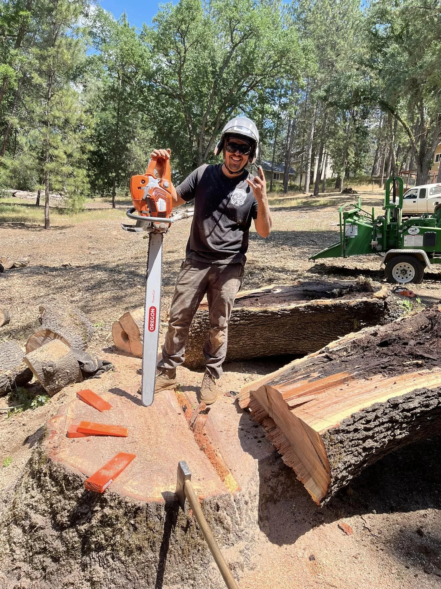 A man standing on a large tree stump holding a chainsaw, making a hand sign, wearing a helmet and sunglasses, in a wooded outdoor area with trees and equipment in the background.