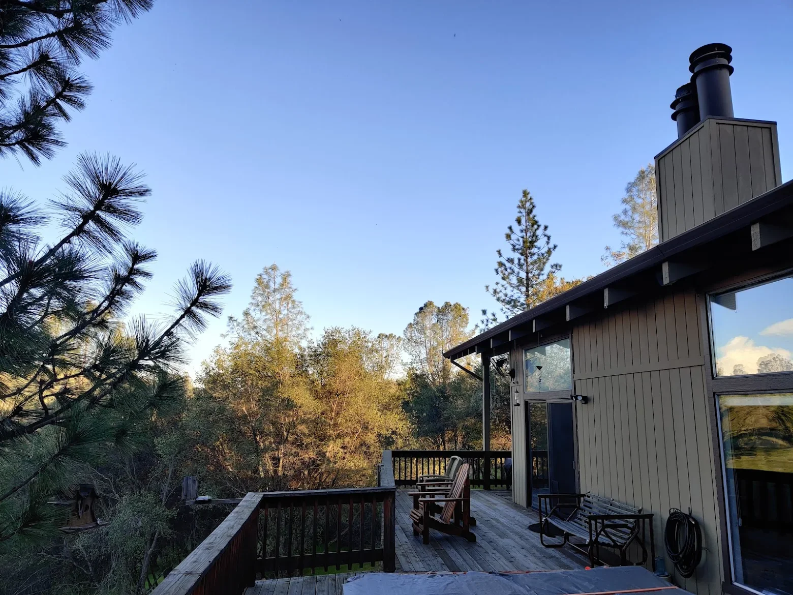 A wooden deck attached to a house with chairs and a garden hose, surrounded by trees and overlooking a forested area under a clear blue sky.