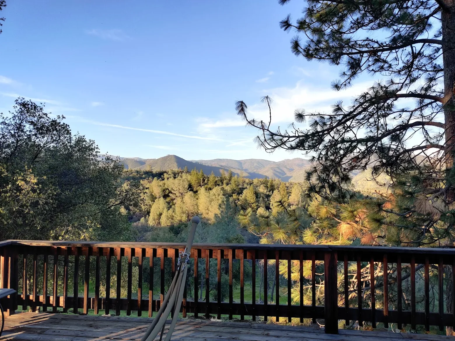 A wooden deck with a railing overlooks a lush green forested landscape with mountain views under a blue sky.