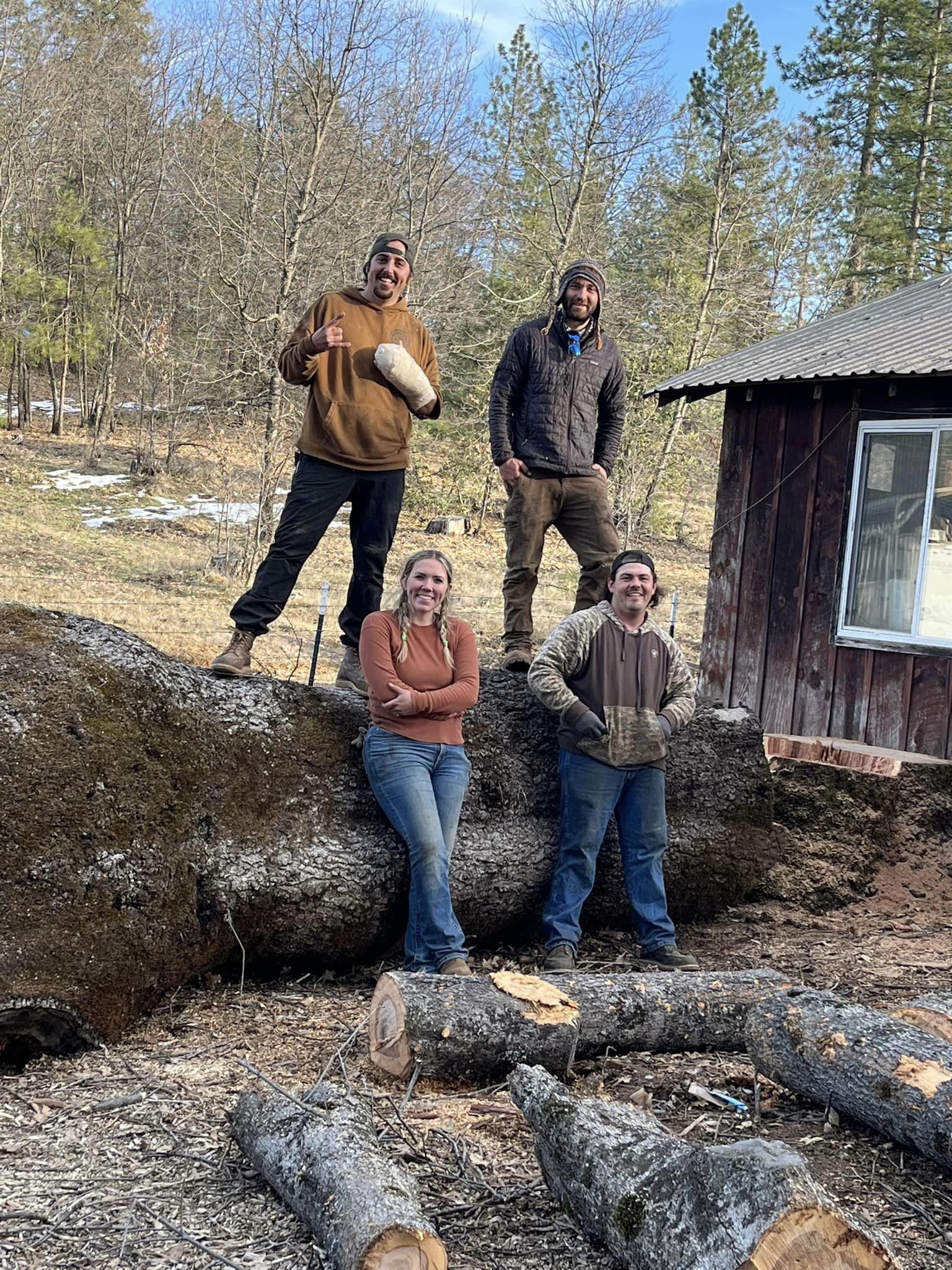 Five people standing outdoors on a fallen log with trees and a wooden shed in the background. They are smiling and dressed in casual outdoor clothing.