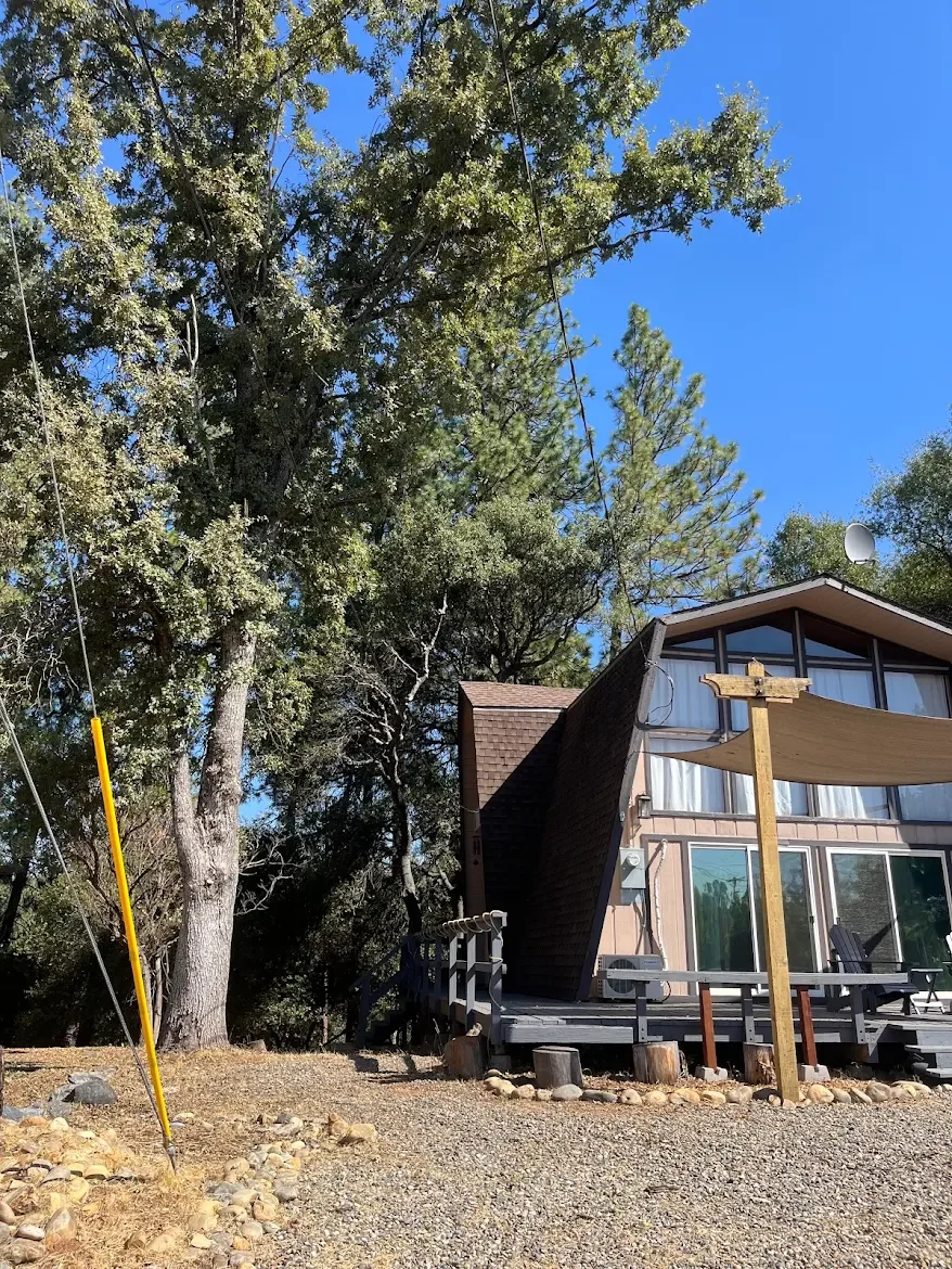 A house with an unusual angular roof, surrounded by tall trees and a bright blue sky. There is a small deck with chairs and a wooden post with a fabric canopy for shade. A clothesline is strung across the front yard, and the ground is covered with gr