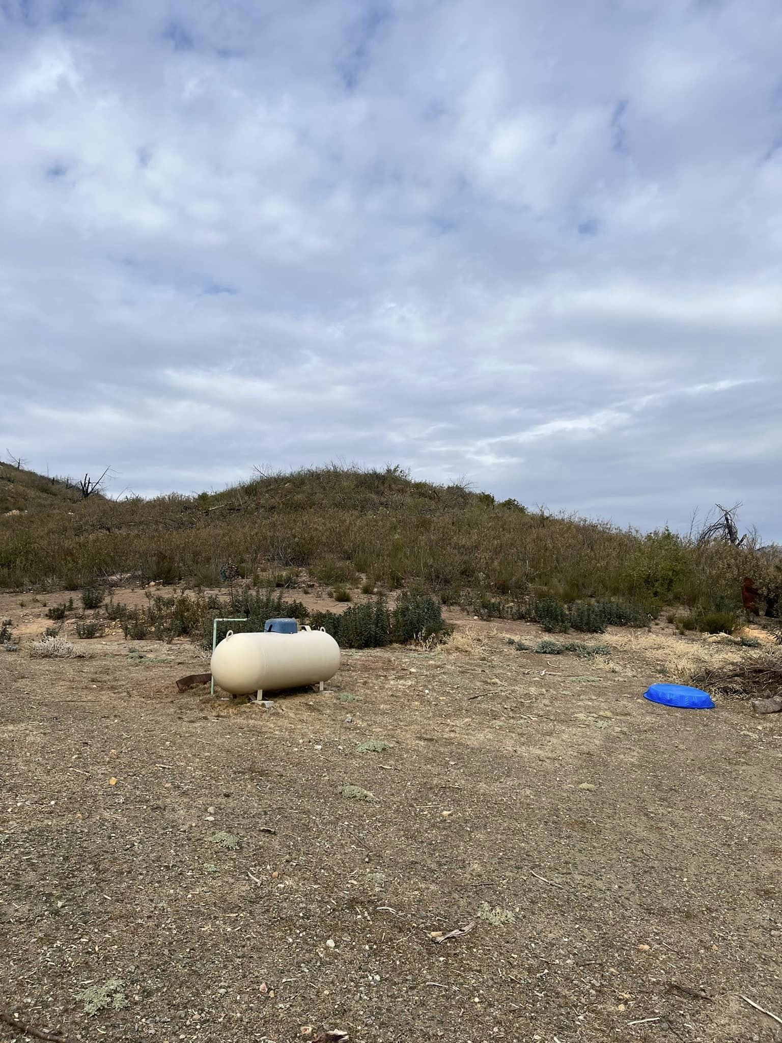 A dry, dirt-covered landscape with sparse shrubbery, a large white propane tank, and a small blue plastic pet or animal water dish, under a cloudy sky.