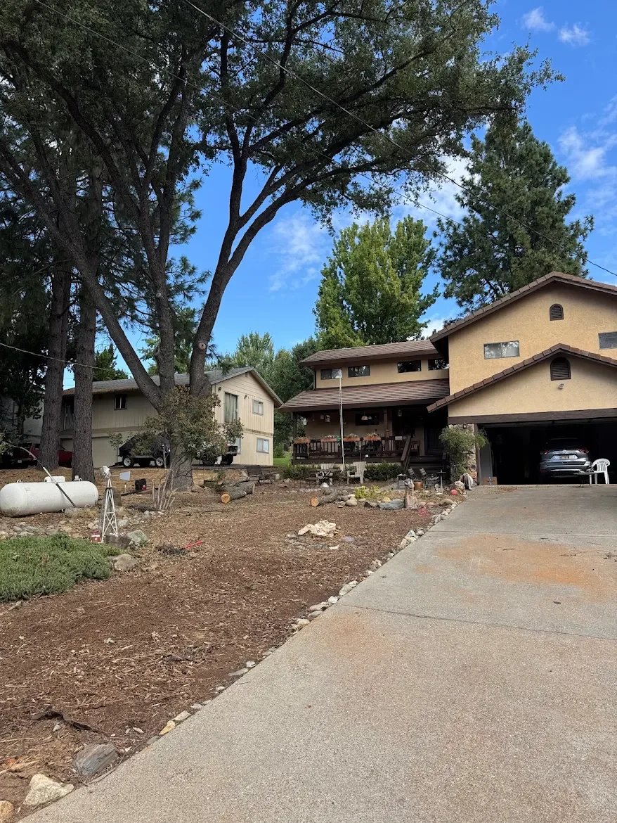 A residential neighborhood with a house on the right and another house across the street, trees, and a driveway. The yard in the foreground appears to be under landscaping or gardening work.