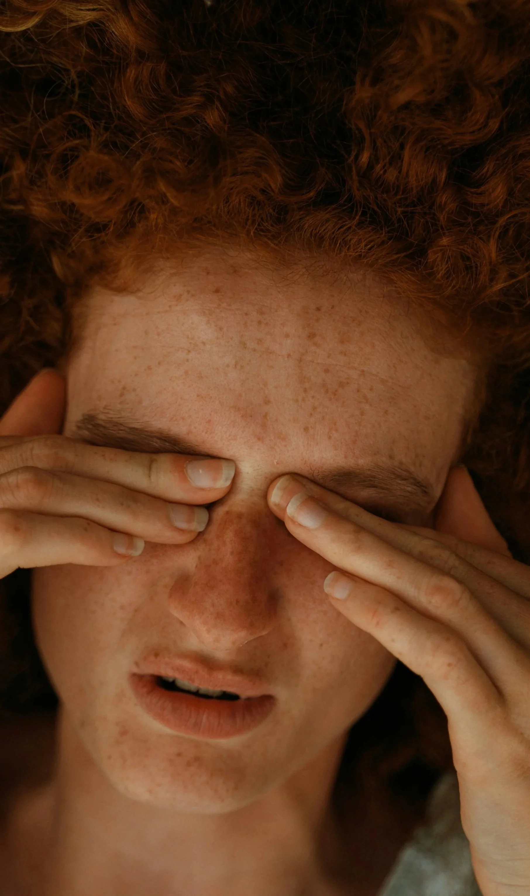 Close-up of a woman with red curly hair holding her face with her hands, eyes closed, with a distressed or tired expression. She has a migraine.