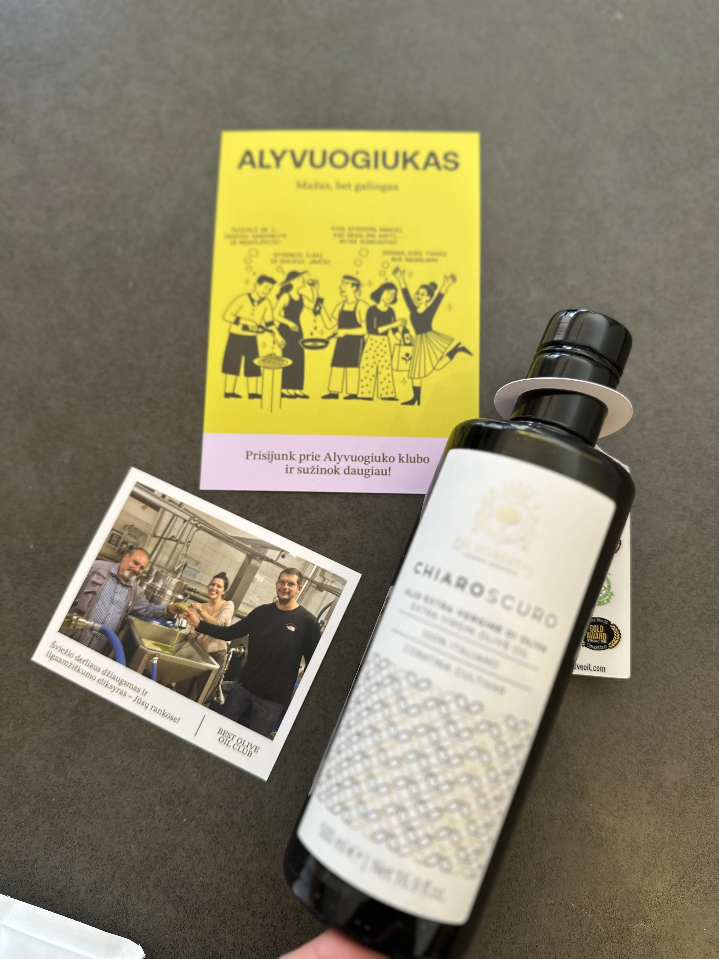 A bottle of Chiaro Scuro extra virgin olive oil with a gold award badge, a yellow flyer promoting an event or club meeting, and a photo of three people in a kitchen, smiling and shaking hands.