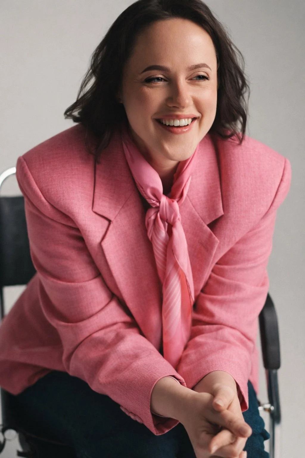 Woman with shoulder-length dark hair smiling, wearing a pink blazer and pink necktie, sitting on a chair with a gray background.