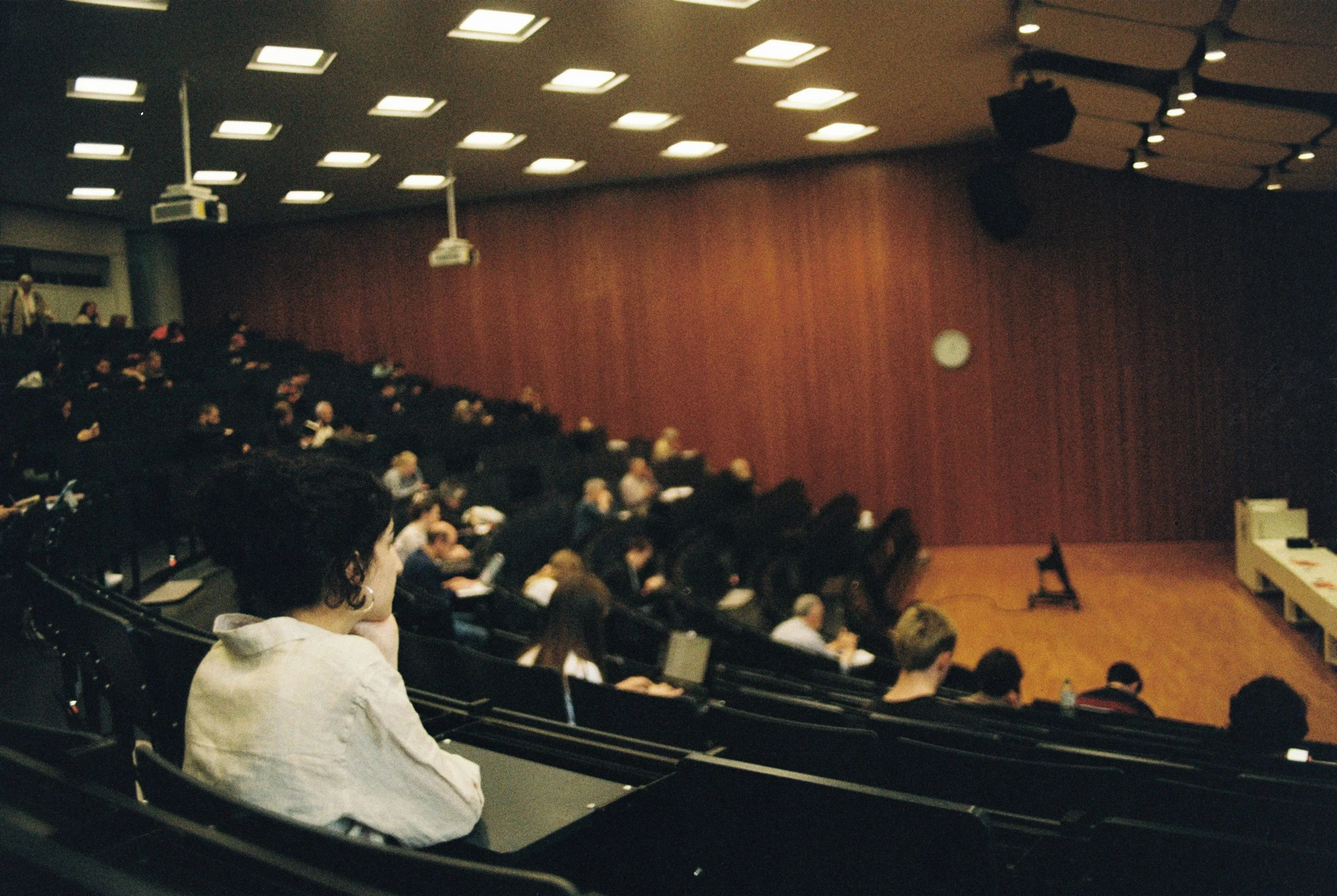 Audience seated in a dimly lit auditorium, facing a stage with a wooden back wall and a clock on the wall. Some people are taking notes or looking at the stage.
