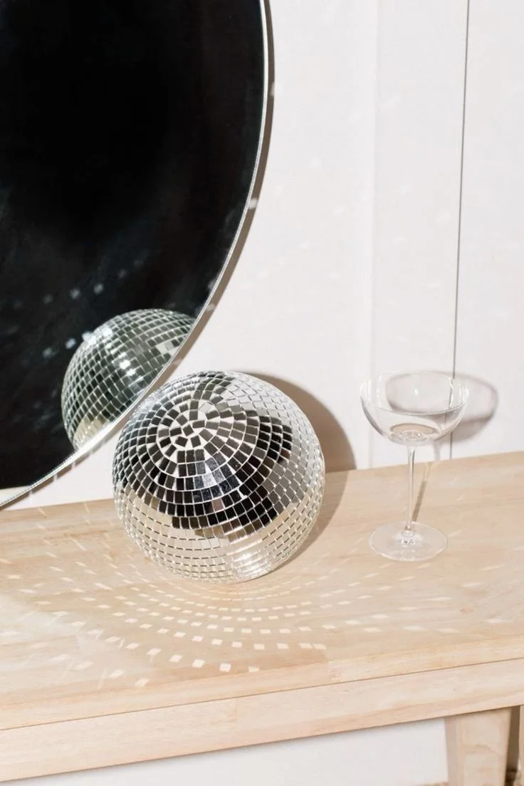 A reflective disco ball, a clear glass bowl on a thin stem, and a mirror are placed on a light wooden surface against a white wall.
