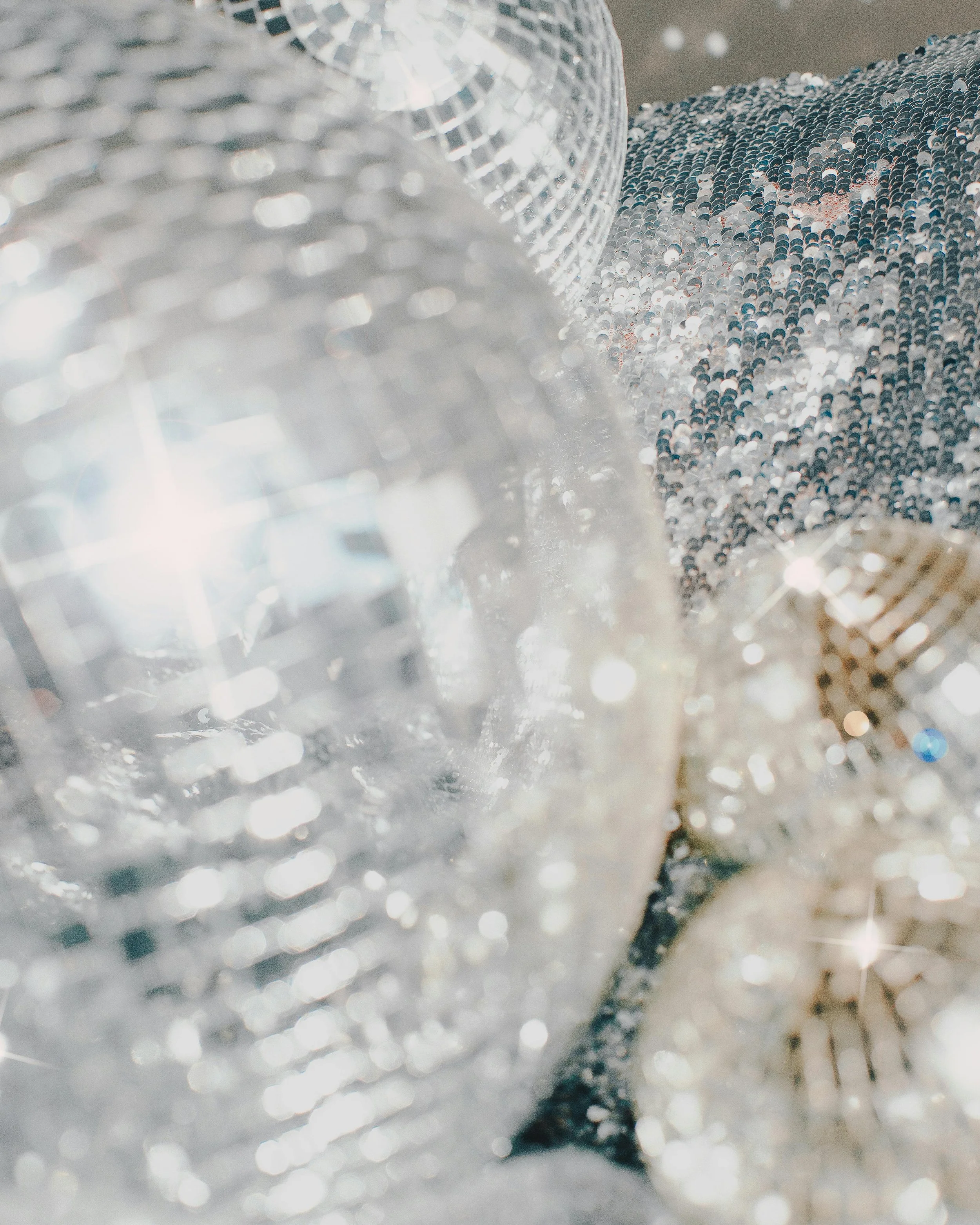 Close-up of a shiny, reflective disco ball with many small mirrored tiles, with another similar disco ball in the background, creating a sparkling and shimmering effect.
