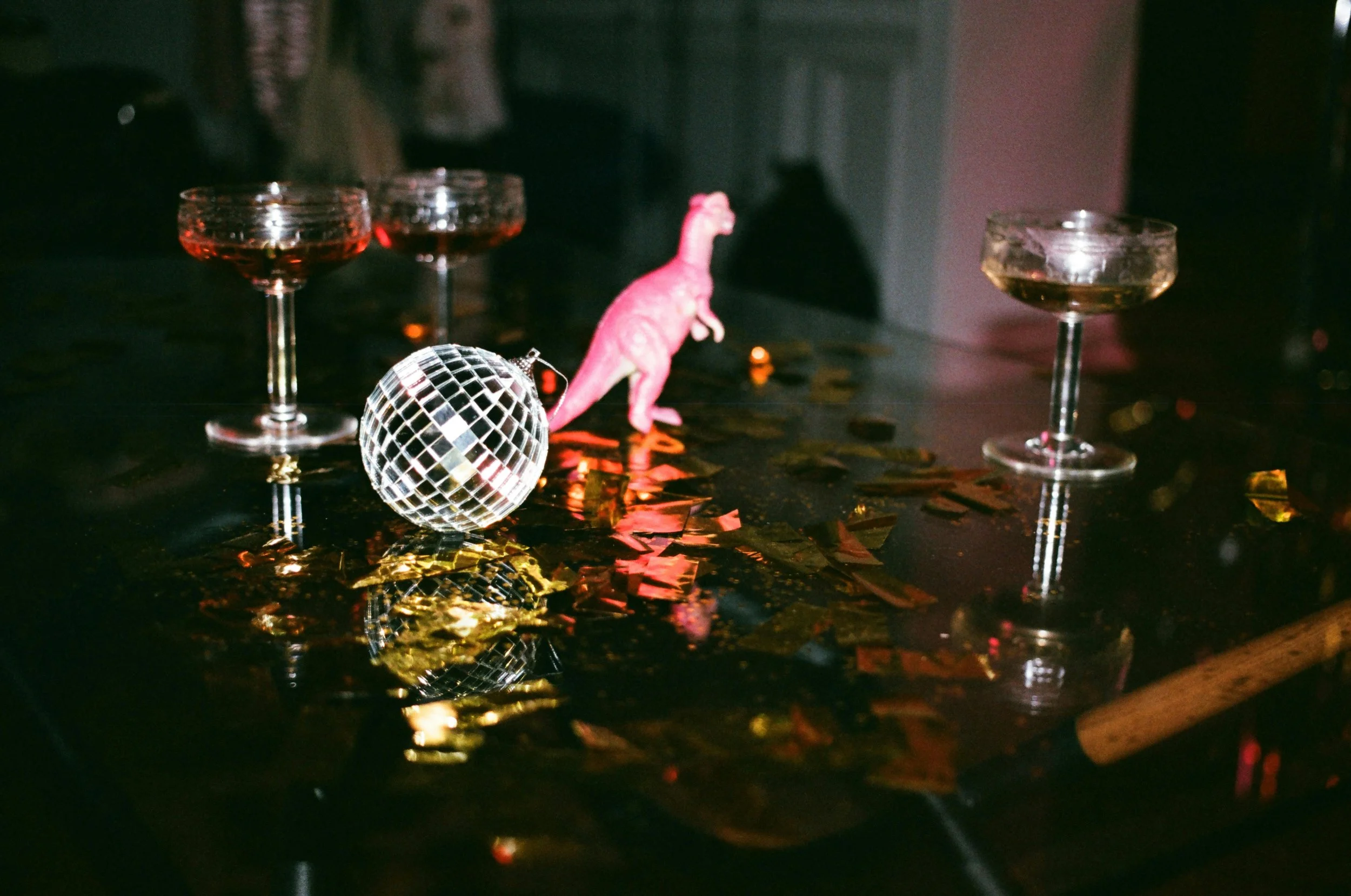 Table with four empty cocktail glasses filled with red drink, a small pink toy dinosaur, a reflective disco ball, and scattered gold and red confetti.