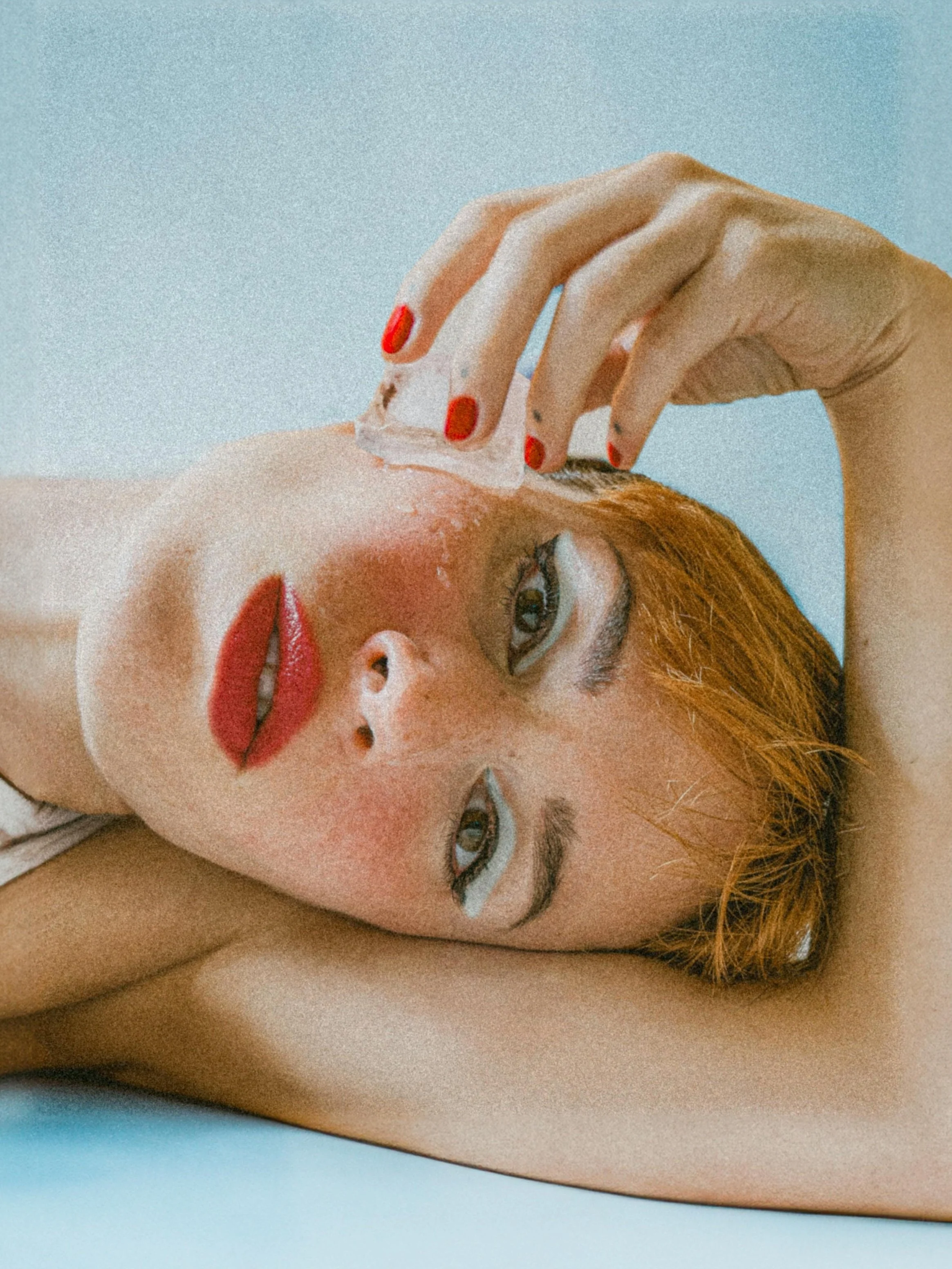 A woman with red hair lying down, with her eyes closed, applying a facial mask with a cotton pad. She has red lipstick and painted fingernails.