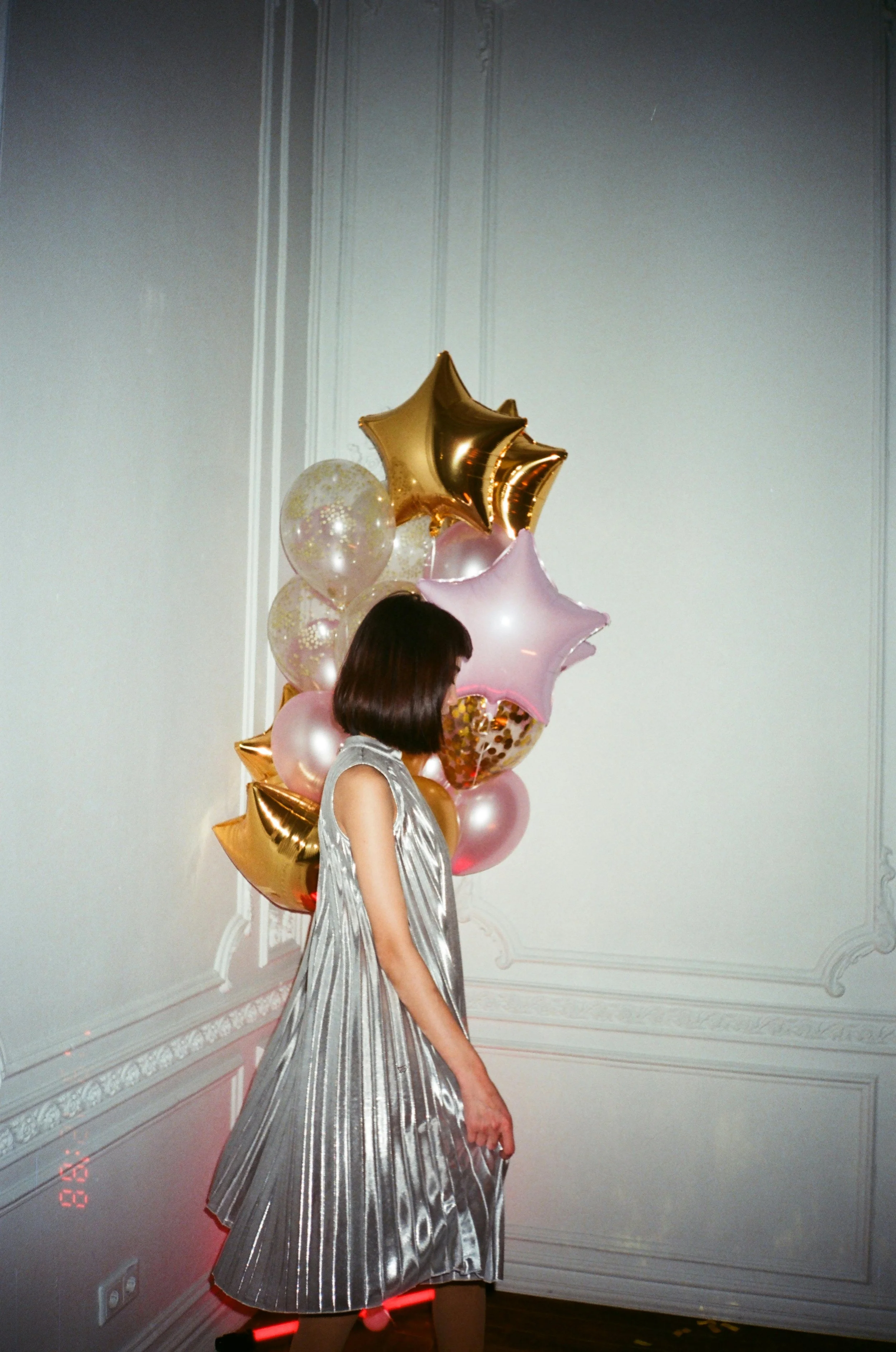 A woman in a shiny silver pleated dress standing in a room with decorative molding, holding a bunch of balloons including star-shaped gold and pink balloons and round balloons with glitter, in front of a plain wall.