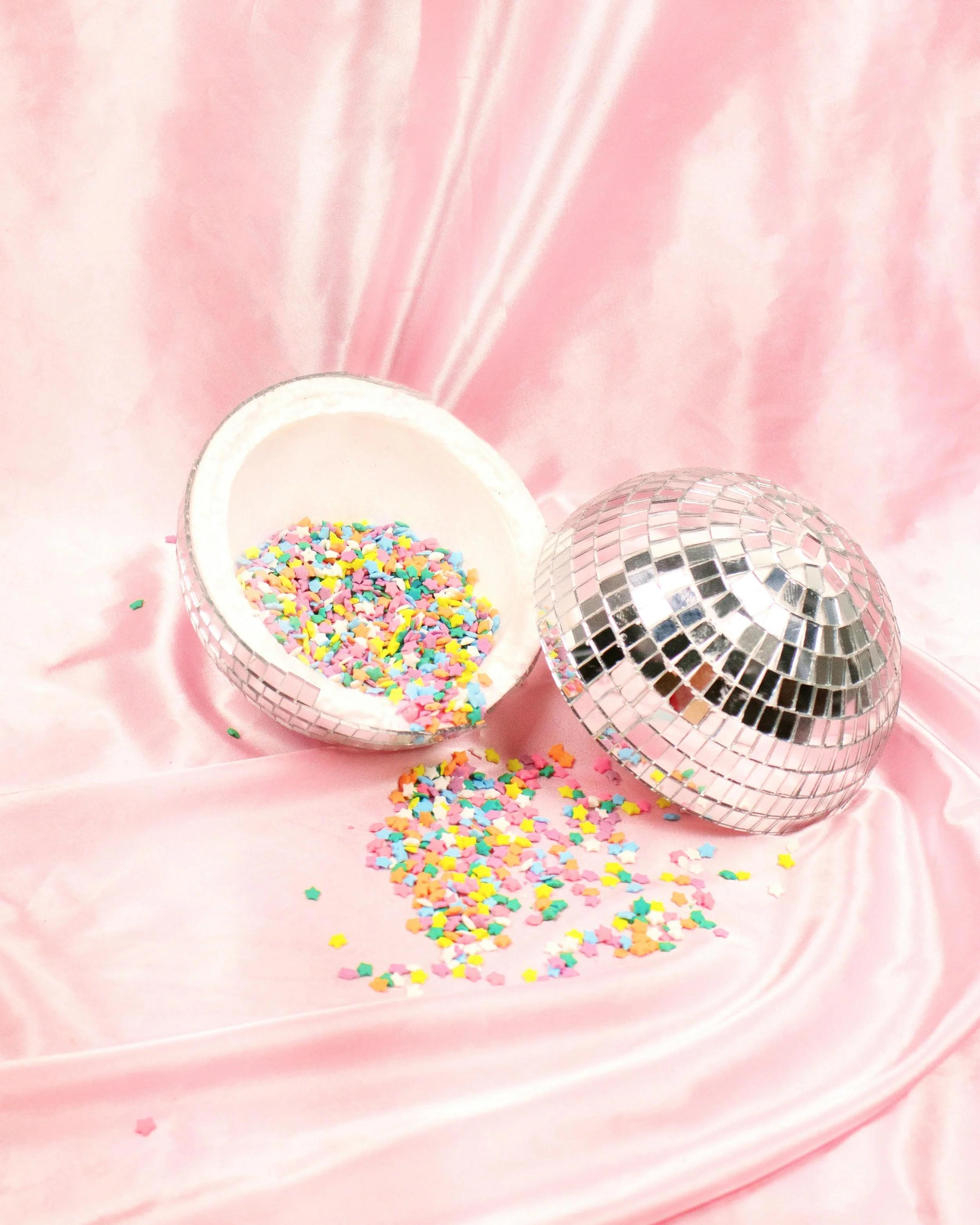 Pink satin backdrop with two mirror disco balls and an open container spilling colorful star-shaped and heart-shaped confetti.