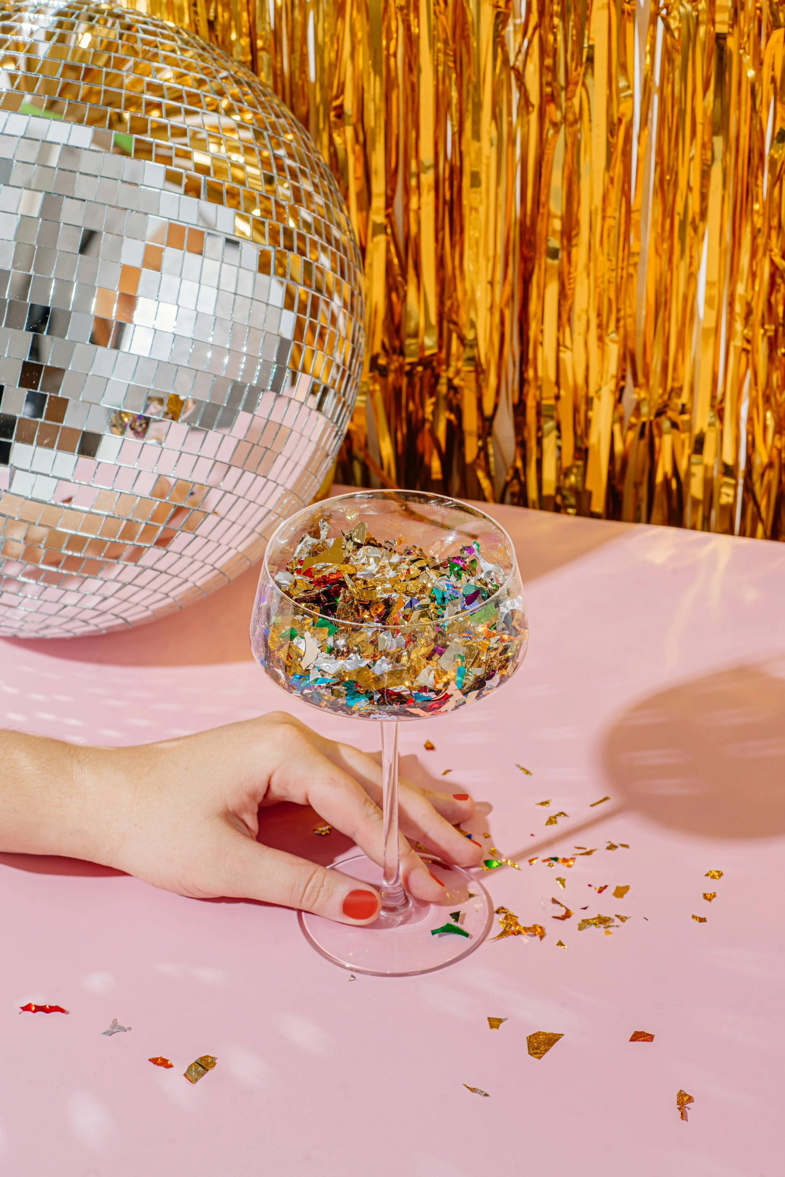 A person with red nail polish holds a clear glass of champagne filled with multicolored confetti, set on a pink table with gold and multicolored confetti scattered around. In the background, there's a disco ball and a gold tinsel backdrop, suggesting a celebration or party environment.