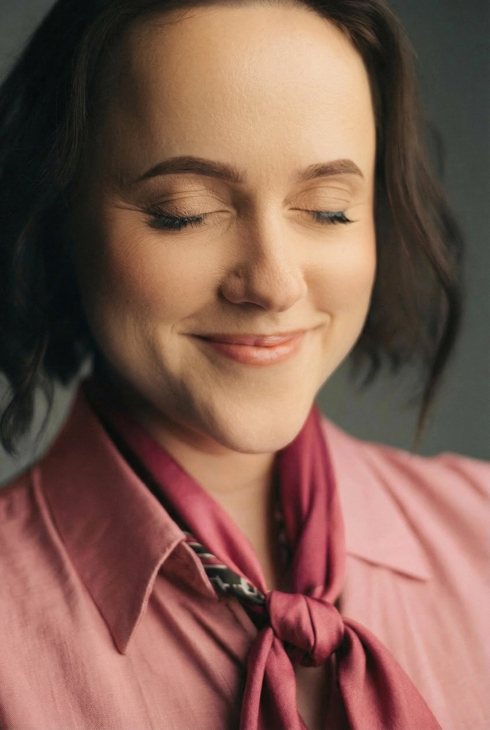 A woman with fair skin and dark brown, wavy hair wearing a pink satin shirt with a matching pink scarf tied around her neck.