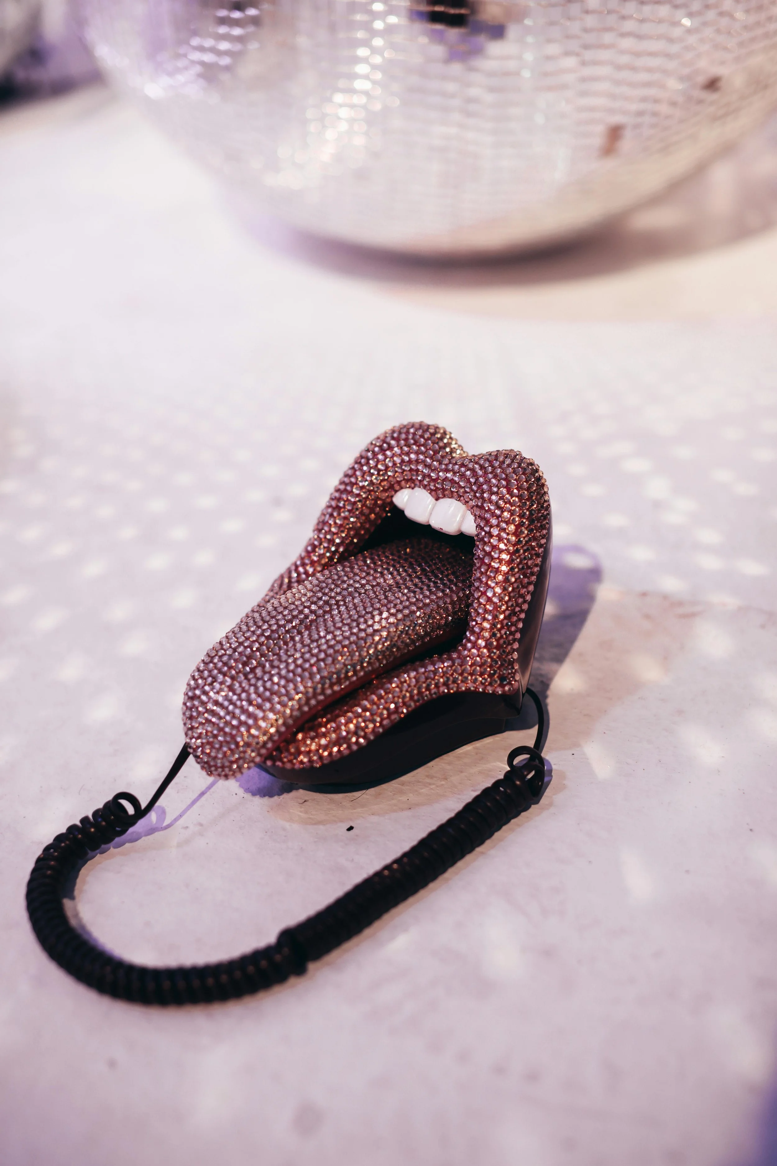 Marketingo ir brandingo paslaugos. A shoe designed to look like lips, covered in pink and purple rhinestones, with teeth detail at the opening, connected to a coiled black cord.