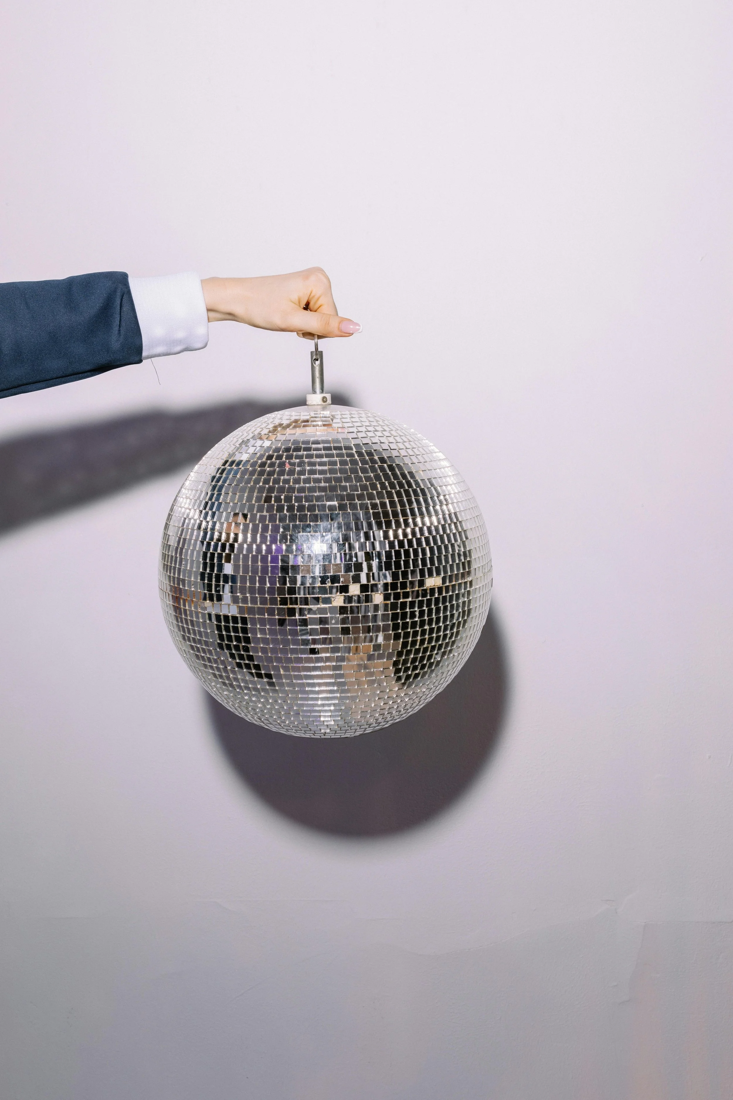 A disco ball hanging from the ceiling with a hand holding a metal hook at the top, casting a shadow on a plain white wall.