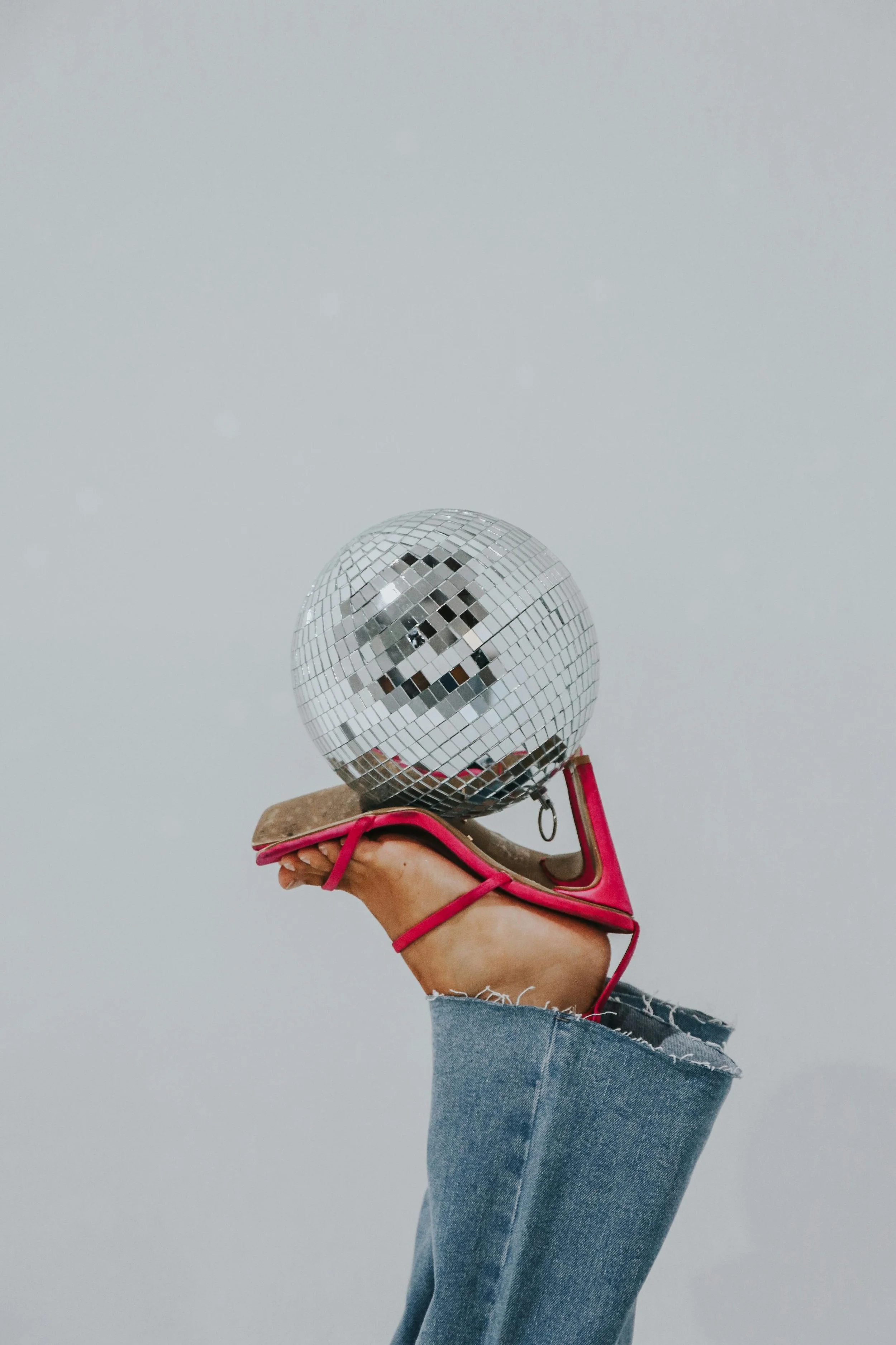 A person's hand holding a disco ball decorated with small mirrored tiles, wearing a pink sandal with a chunky heel, and ripped blue jeans against a plain gray background.
