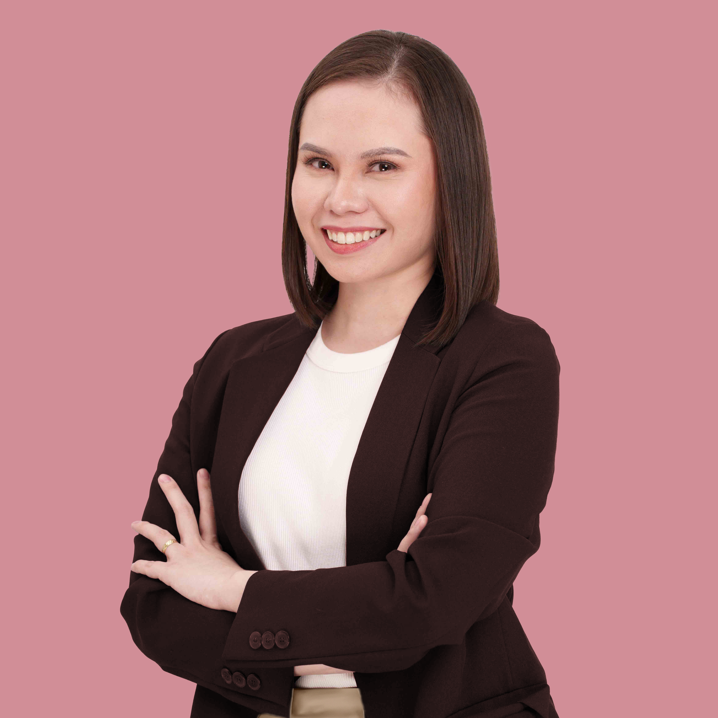 Professional woman with straight brown hair wearing a dark blazer and white top, smiling with arms crossed, against a pink background.