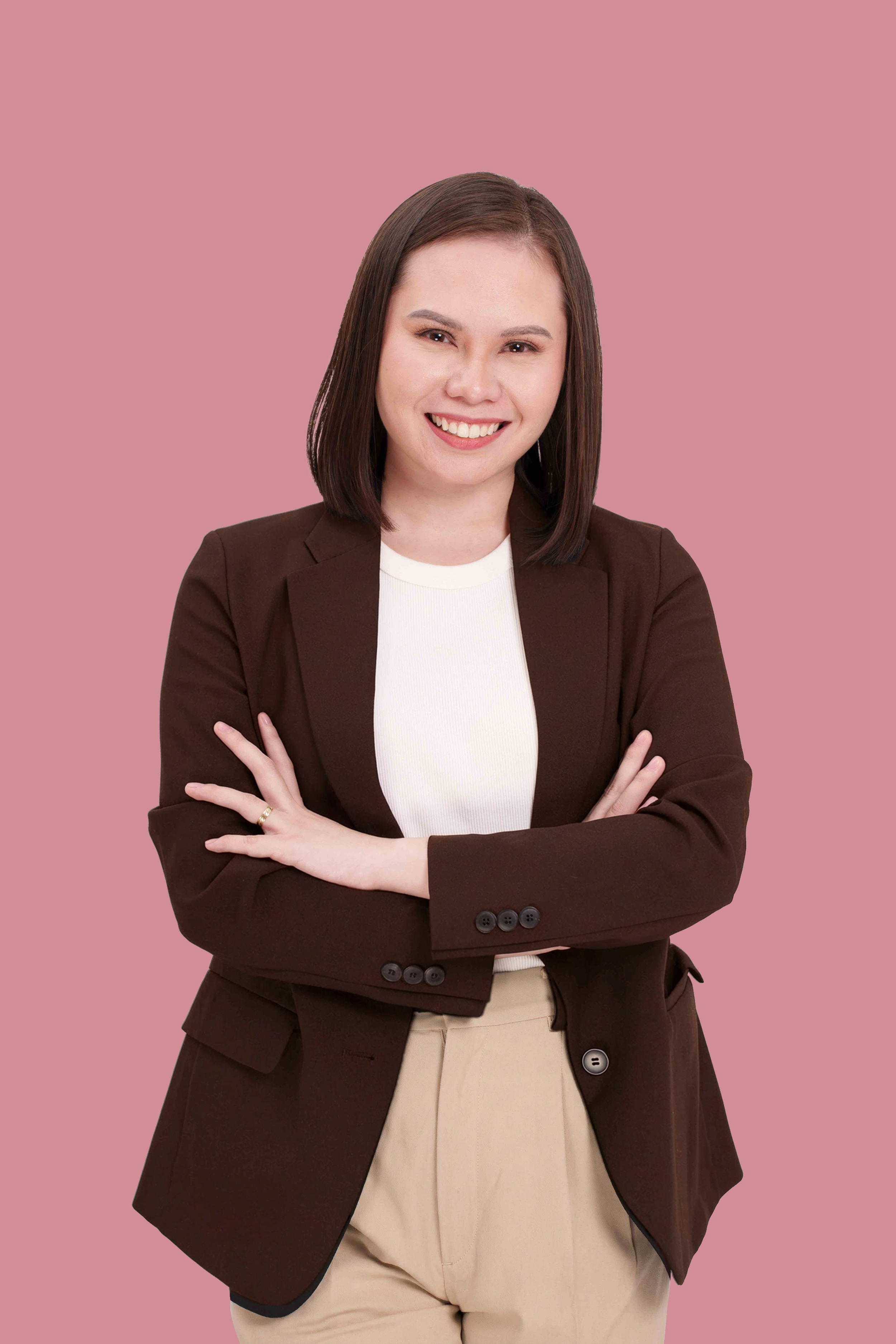 A woman with shoulder-length dark brown hair, wearing a brown blazer over a white top, smiling with folded arms against a pink background.