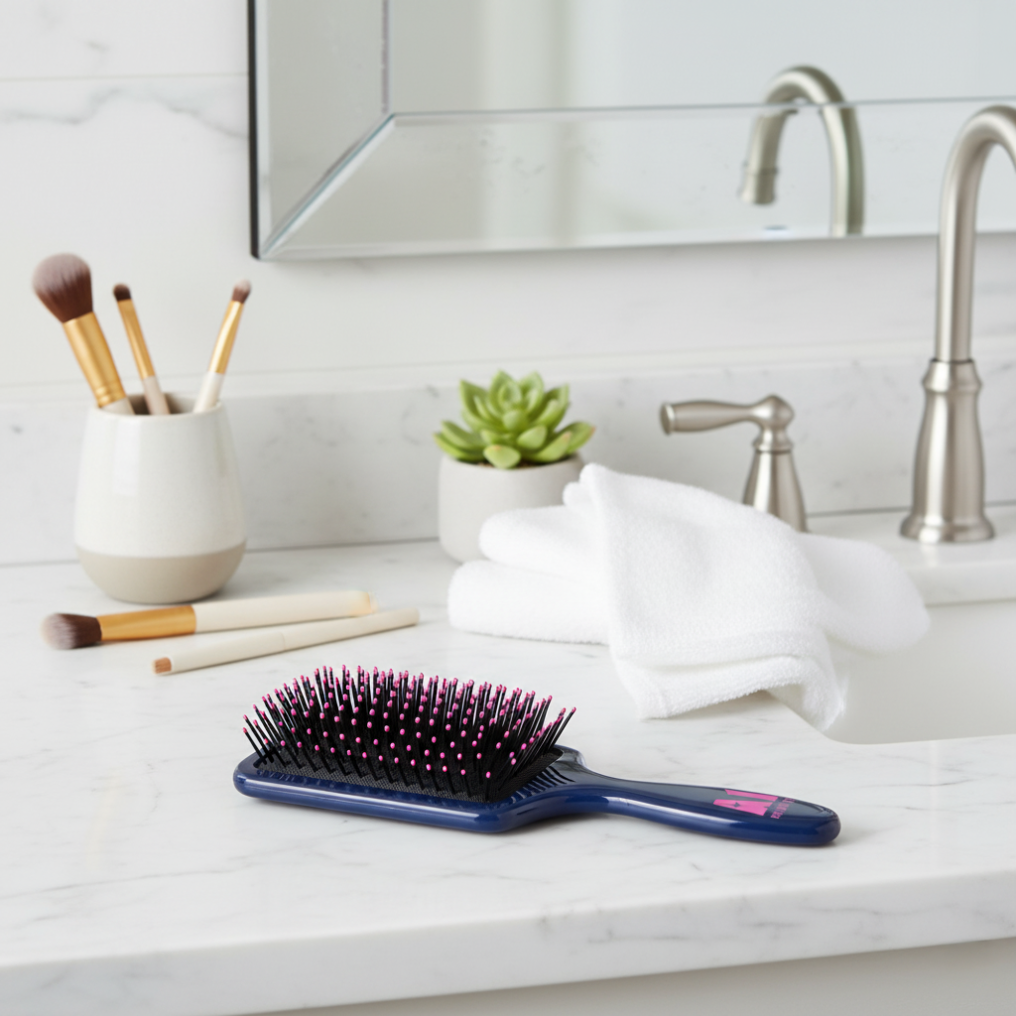 A bathroom counter with a black hairbrush with pink and black bristles, a white towel, a small green succulent plant in a white pot, and a white ceramic container holding makeup brushes, with a mirror and a metallic faucet in the background.