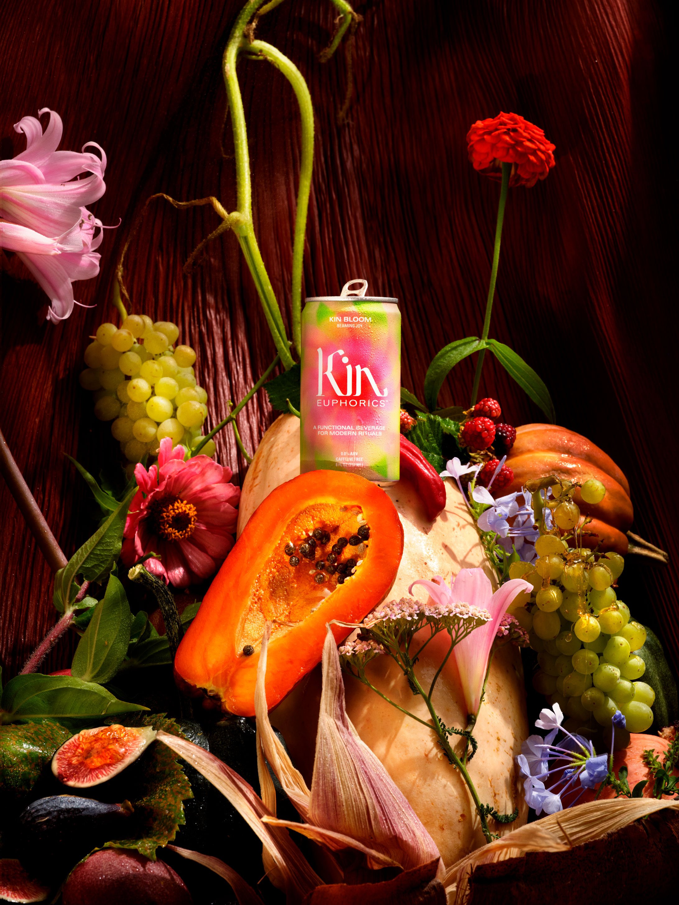 Arrangement of fruits, flowers, and a Kin Euphorics drink can on a wooden surface, including papaya, grapes, berries, figs, and various colorful blooms.