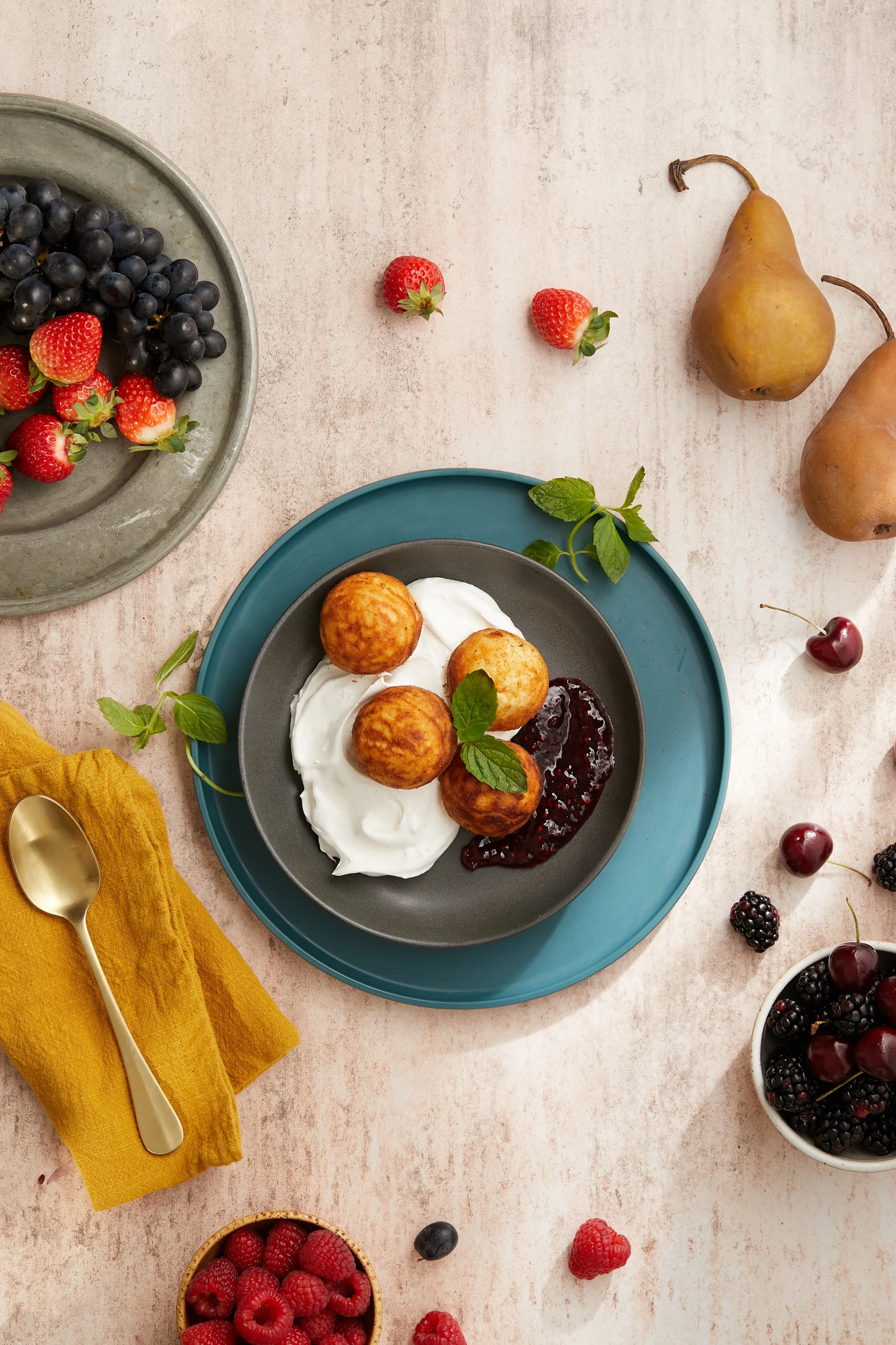 A black plate with four golden brown fried dough balls, served with yogurt and berry jam, garnished with fresh mint leaves, surrounded by bowls of fresh strawberries, blackberries, cherries, and raspberries, on a textured light pink background with a yellow napkin and gold spoon.