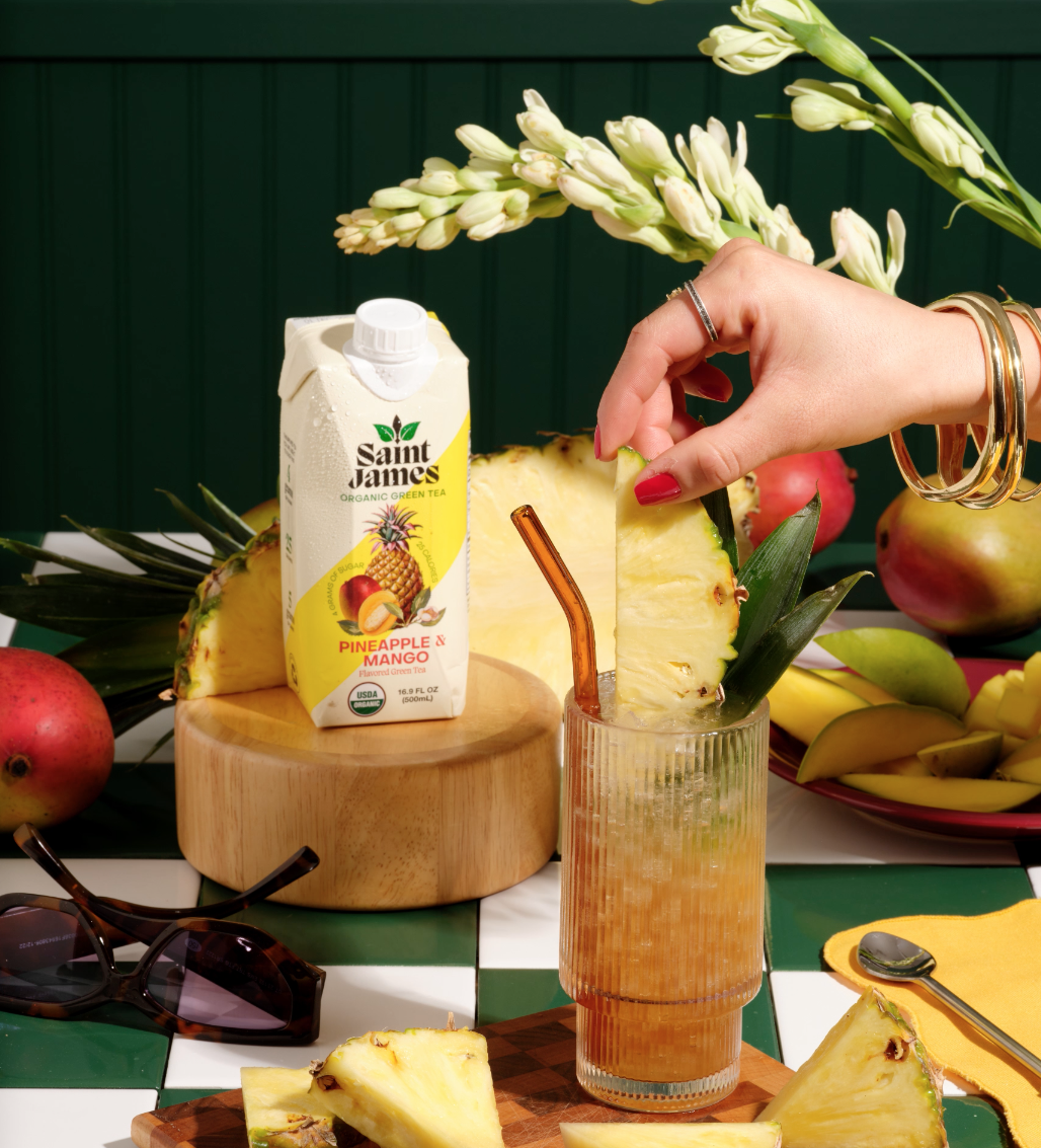 A woman’s hand with red nails and bracelets holding a pineapple slice over a glass of pineapple juice, with tropical fruits, a carton of Saint James pineapple and mango organic green tea, sunglasses, and floral decorations on a checkered green and white table.