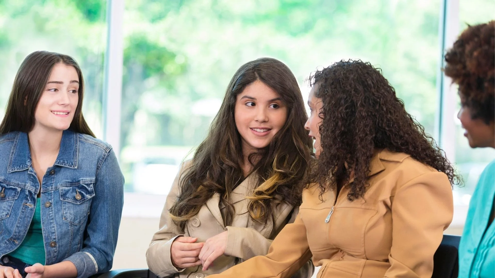 Four women sitting in a circle, engaging in conversation indoors with a large window showing greenery outside.