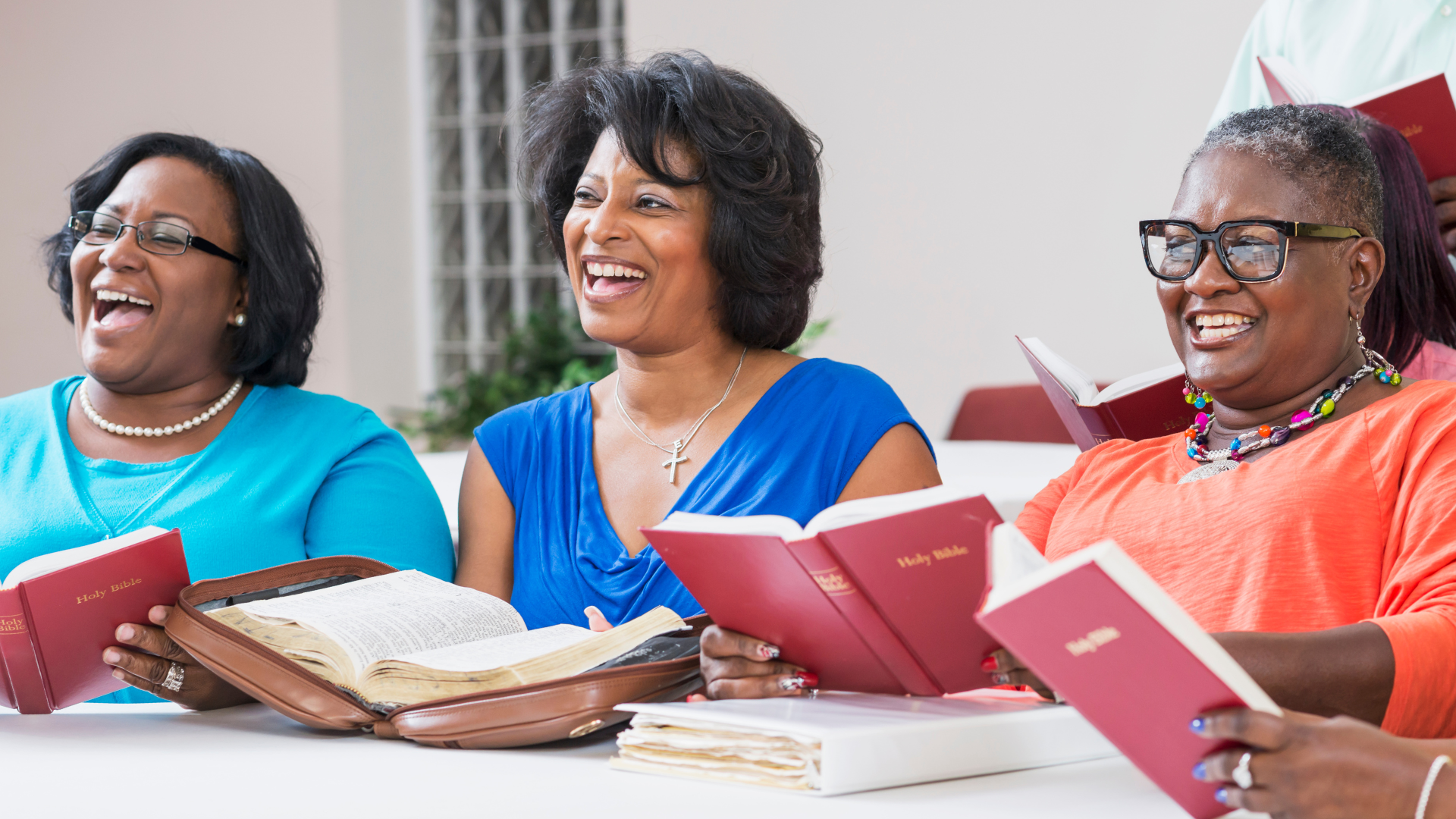 A group of women sitting together at a table, holding red hymn books, and smiling during a religious gathering or church service.