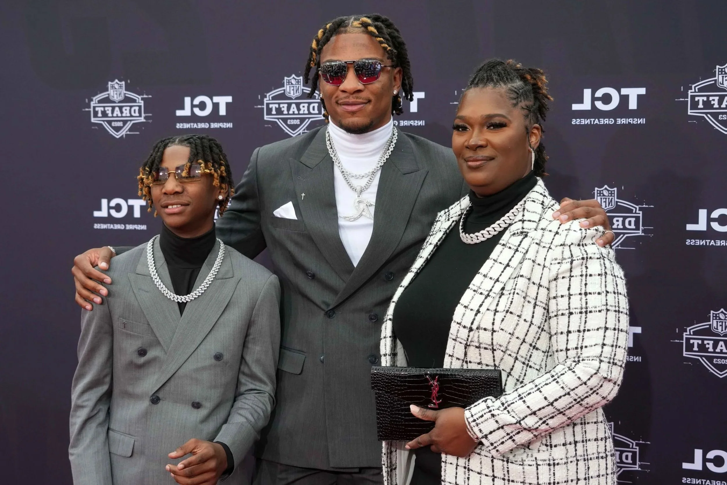 A family of three at an event standing in front of a backdrop with NFL and draft logos. The young man on the left is wearing glasses, a gray suit, and a pearl necklace. The man in the middle is wearing sunglasses, a gray suit, and jewelry. The woman on the right is wearing a plaid blazer, black top, and holding a black clutch. All three are smiling and posing for the photo.