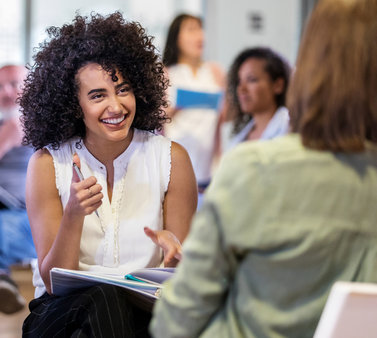 A young woman with curly hair smiling and talking to an older woman in an office or classroom setting.