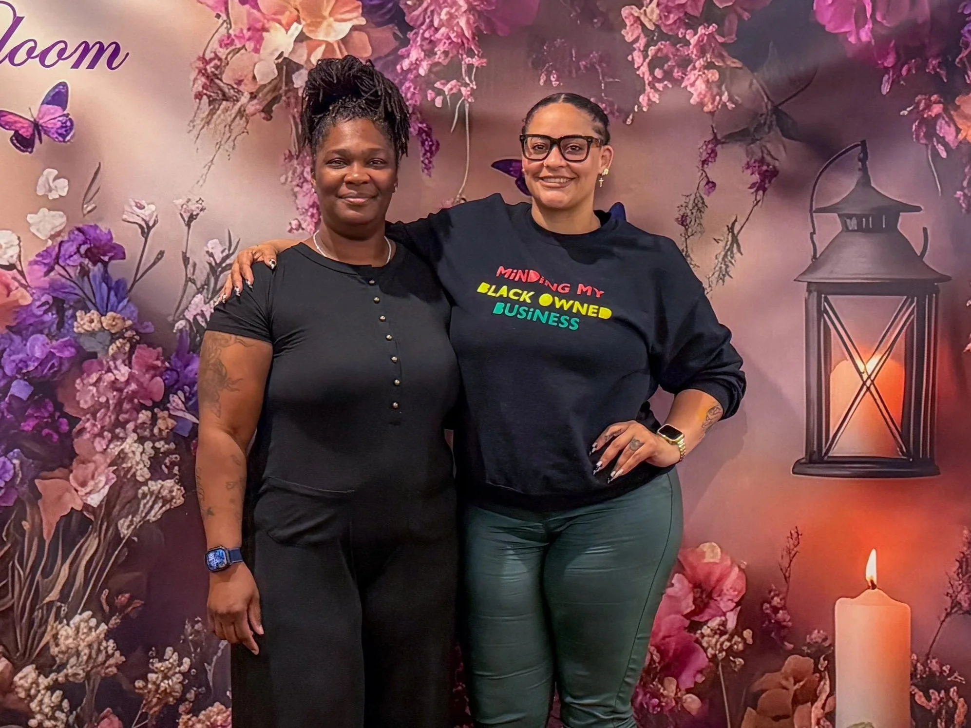 Two women standing together in front of a floral background with a lantern and candle, one with arm around the other's shoulder, smiling at the camera.