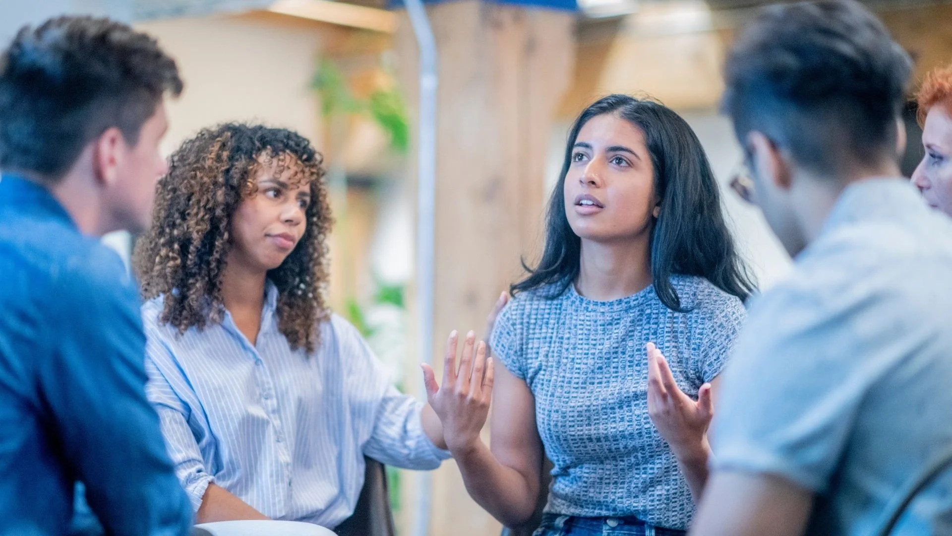 A group of five young adults having a discussion, with one woman speaking and others listening attentively in a casual setting.