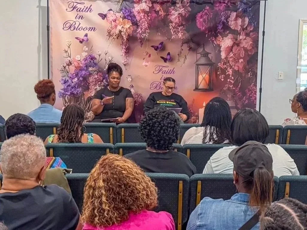 Two women are sitting at the front of a room, presenting or speaking to an audience of diverse adults seated in rows. The backdrop features pink flowers, butterflies, a lantern, and the words "Faith in Bloom."