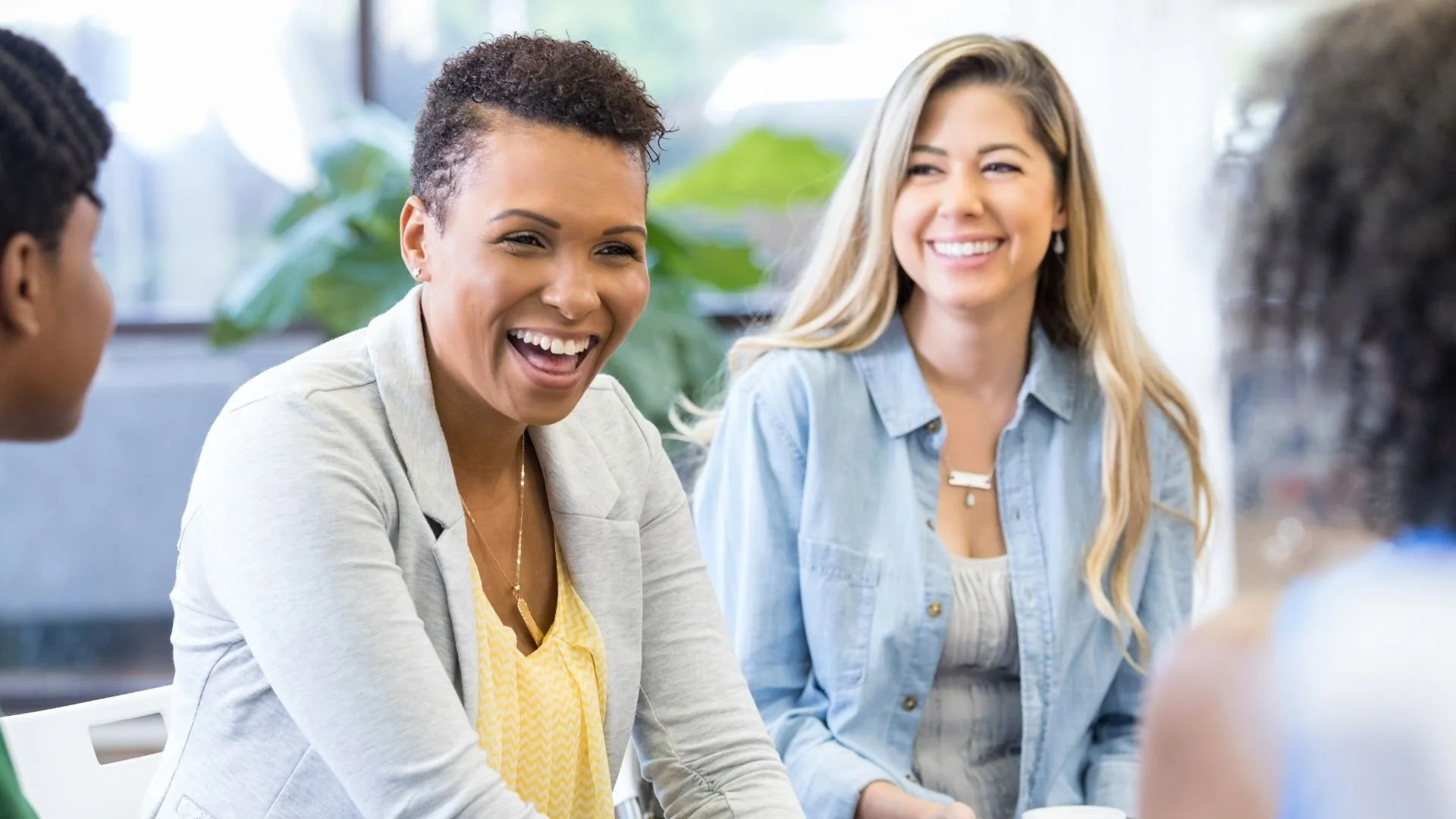 Four women sitting together, engaged in a conversation, smiling, in a bright indoor setting with green plants in the background.