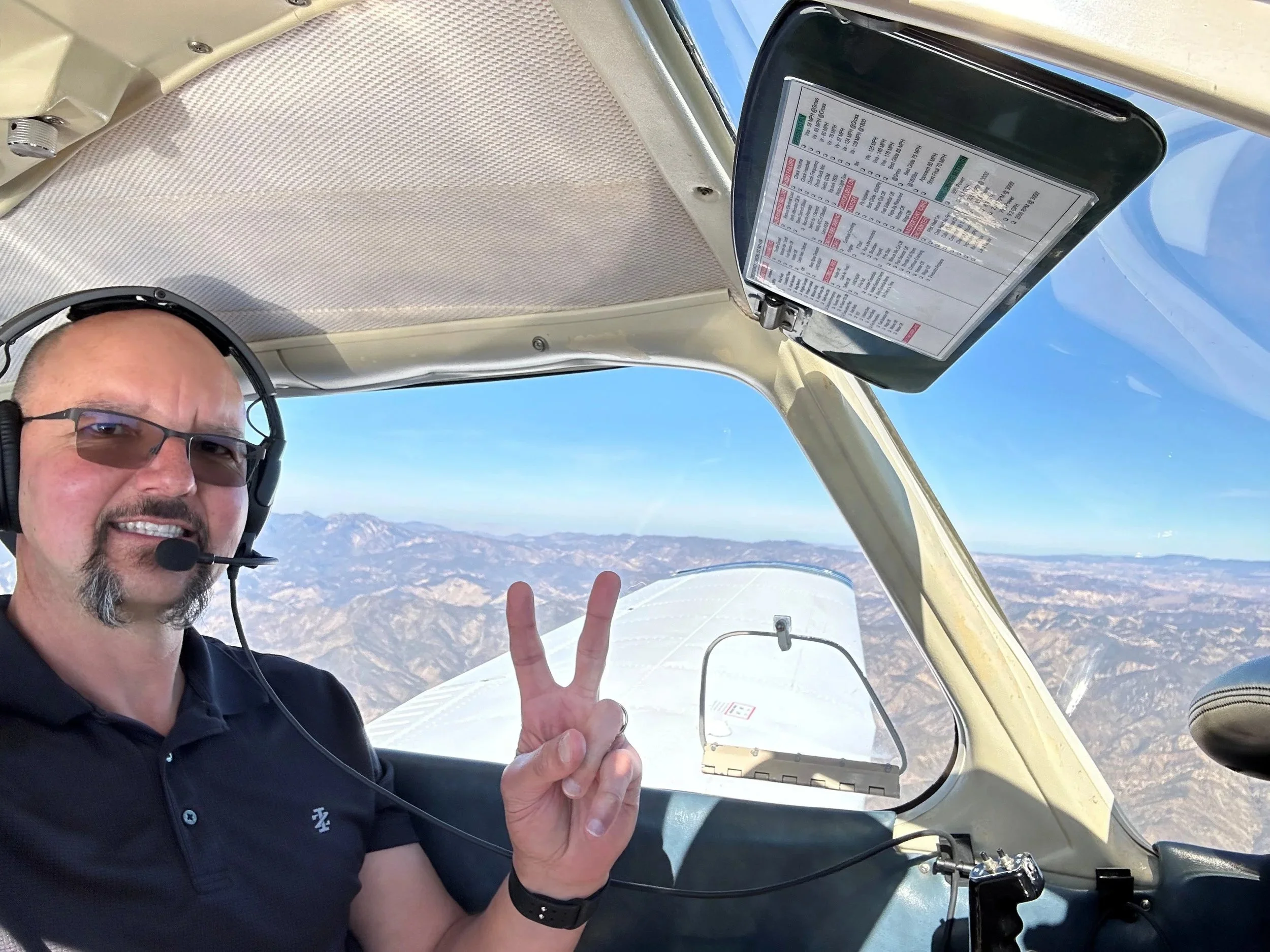 A pilot in the cockpit of a small airplane, smiling and making a peace sign with his hand. The background shows mountainous terrain and a clear blue sky.