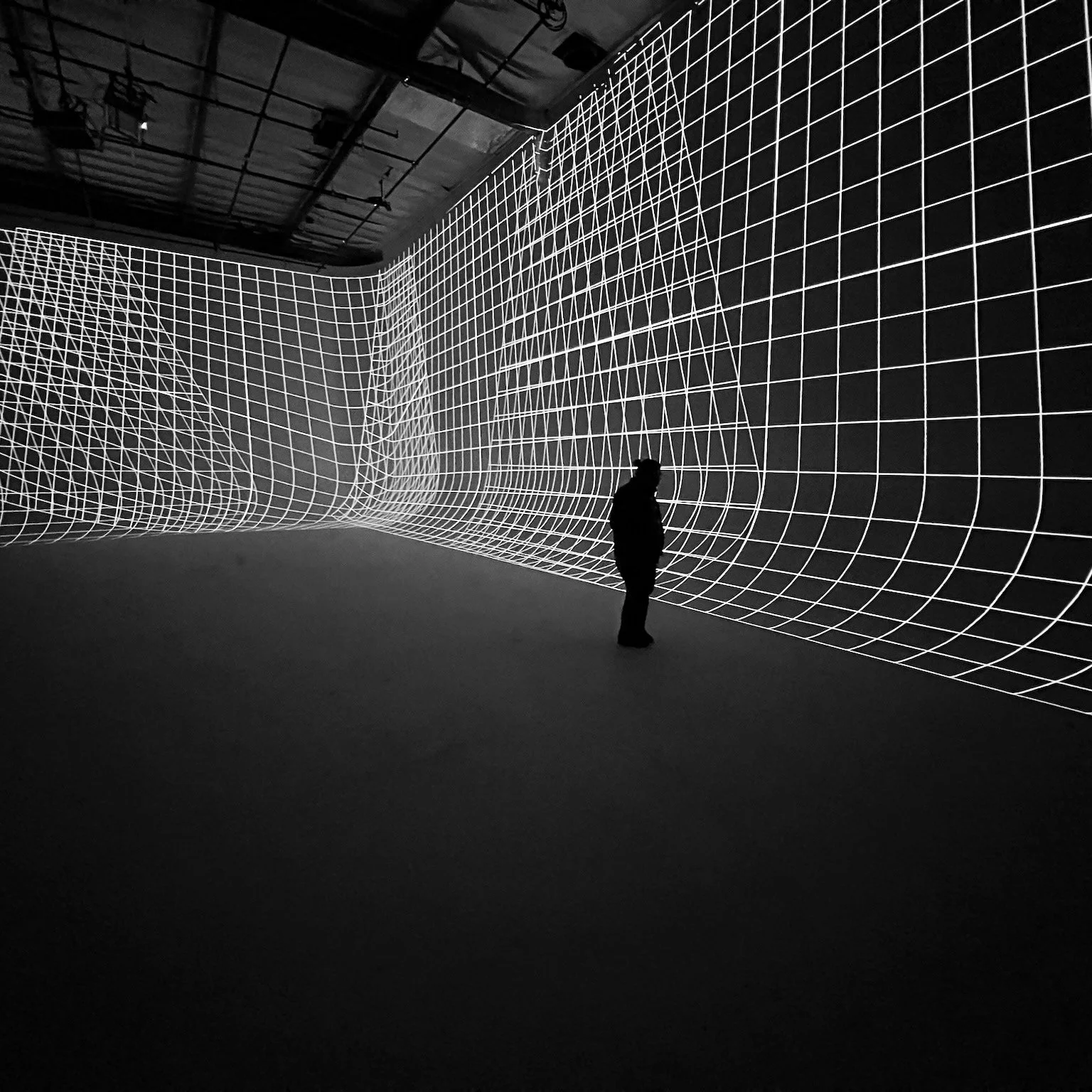 Person standing in an art installation with black walls and white grid lines creating a 3D geometric pattern.
