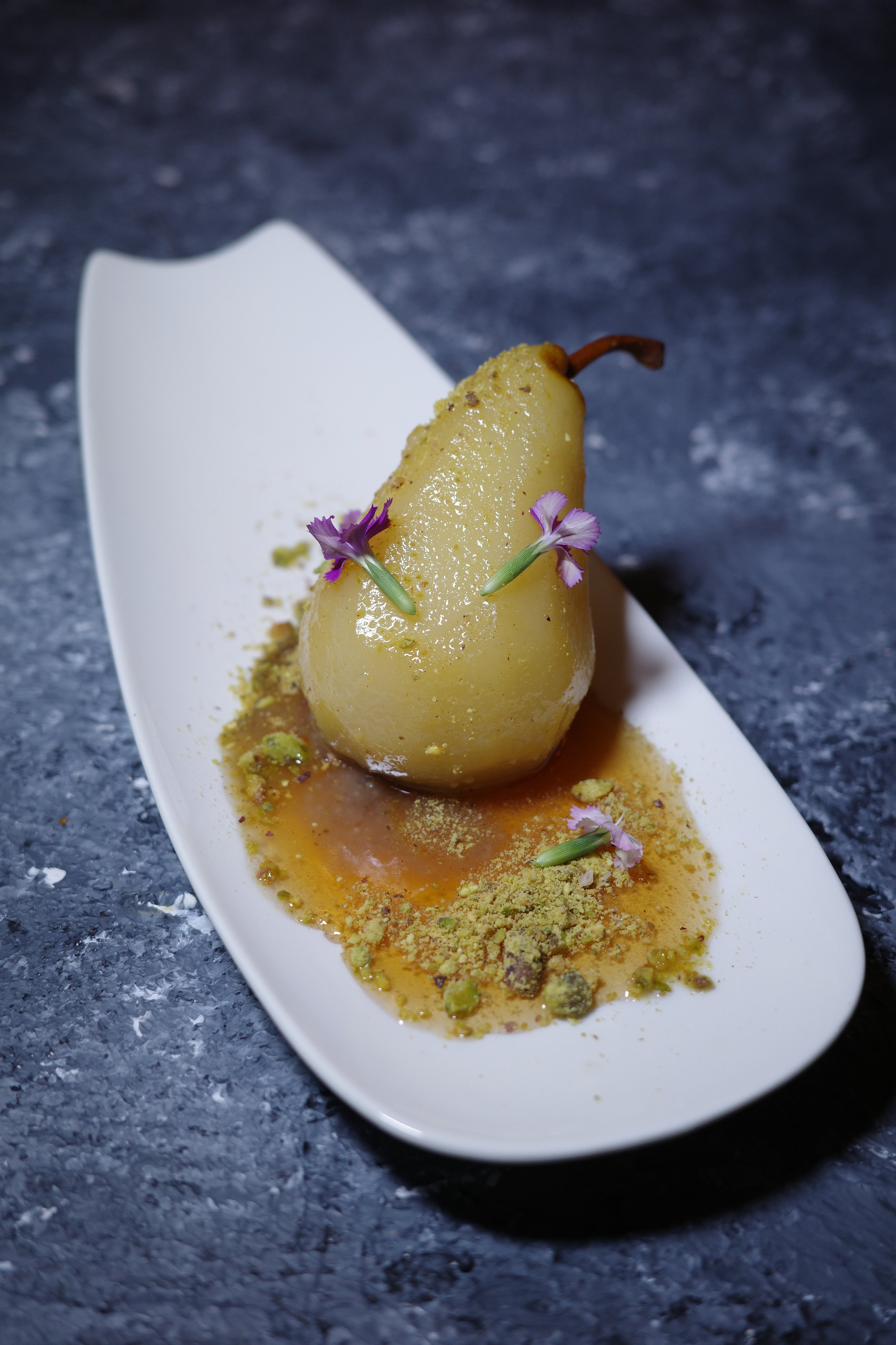 A poached pear garnished with edible flowers and surrounded by crushed pistachios and sauce on a white dish.