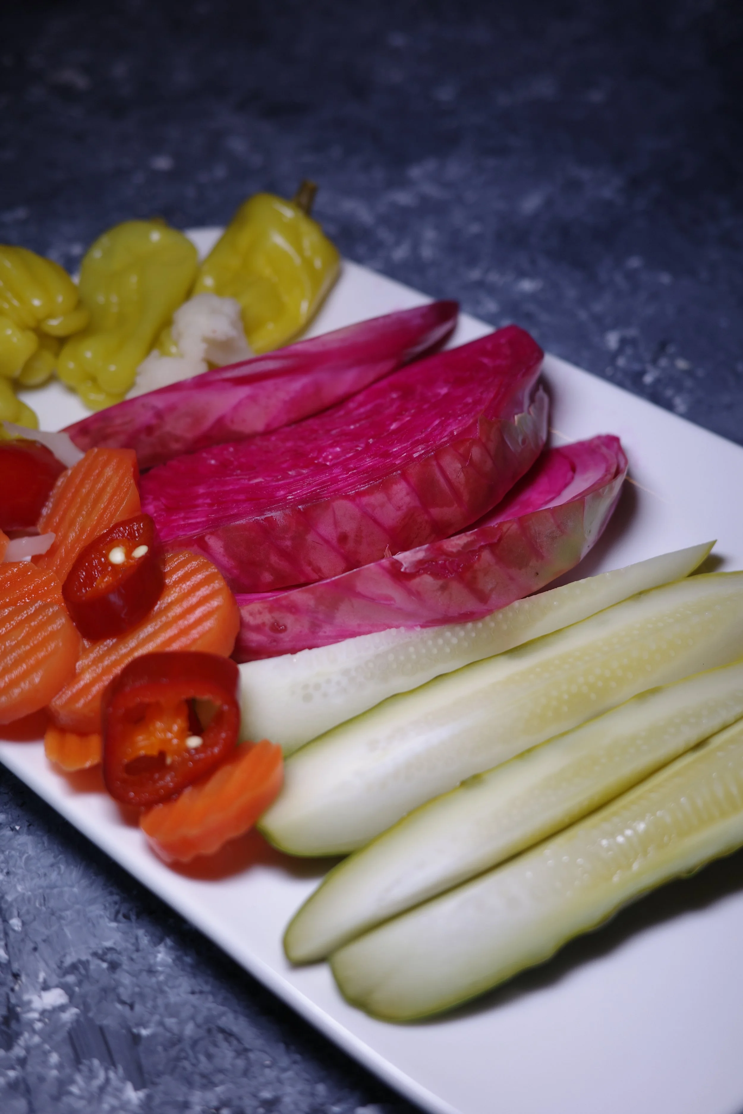 Assorted sliced vegetables on a white rectangular plate, including yellow pickled peppers, pink jicama, orange carrots with red chili slices, and pale green cucumber spears, on a dark textured surface.
