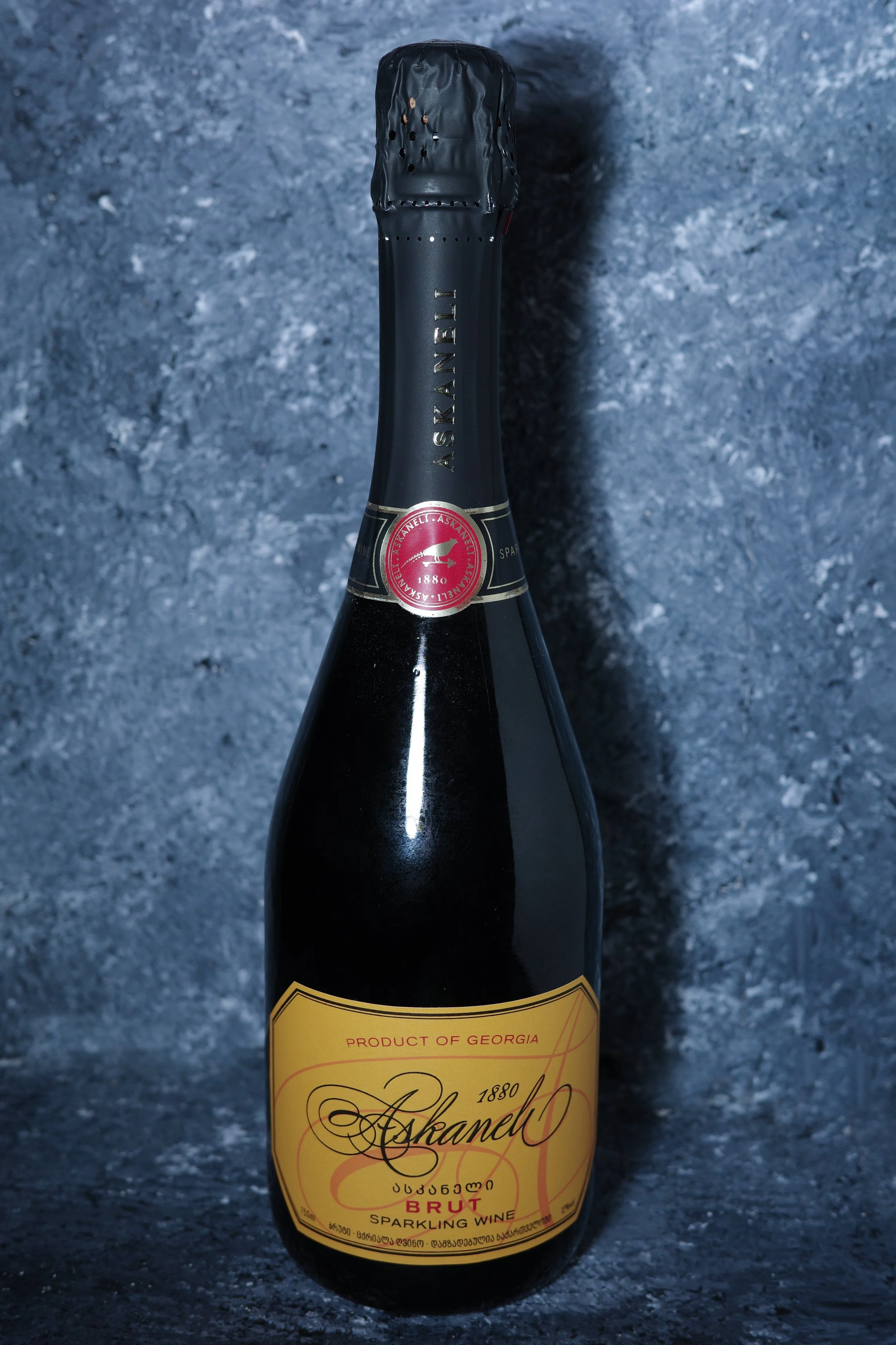 A bottle of Ashanel sparkling wine with a black foil top and a yellow label, set against a textured dark background.