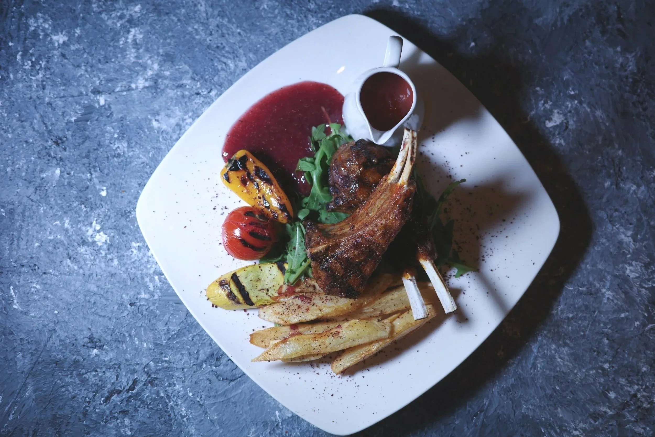 A white square plate with grilled lamb chops, roasted vegetables including cherry tomato, grilled yellow pepper, and potato wedges, garnished with greens, with a small pitcher of red sauce and a dollop of sauce nearby, on a dark textured surface.