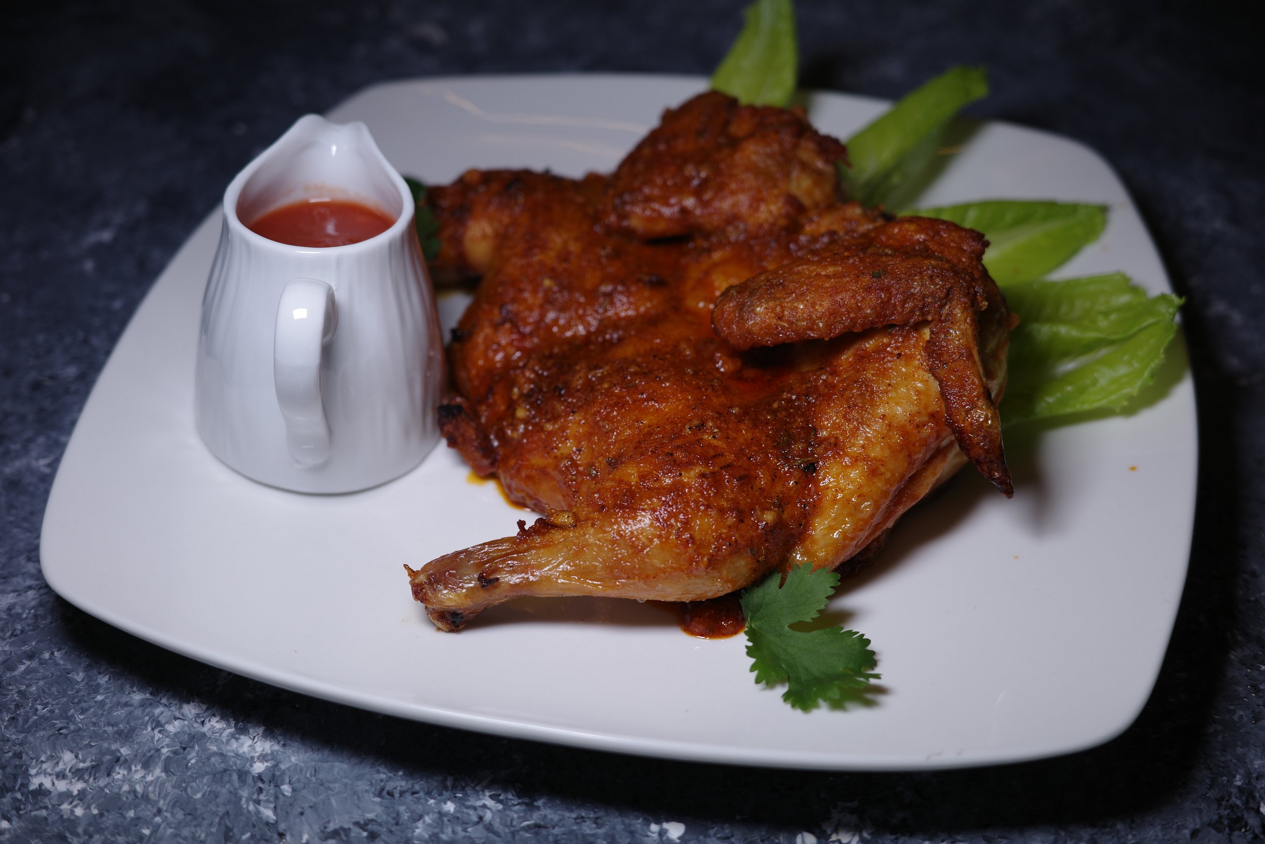 Plate with a cooked chicken leg, lettuce, parsley, and a small white pitcher of red sauce on a dark surface.