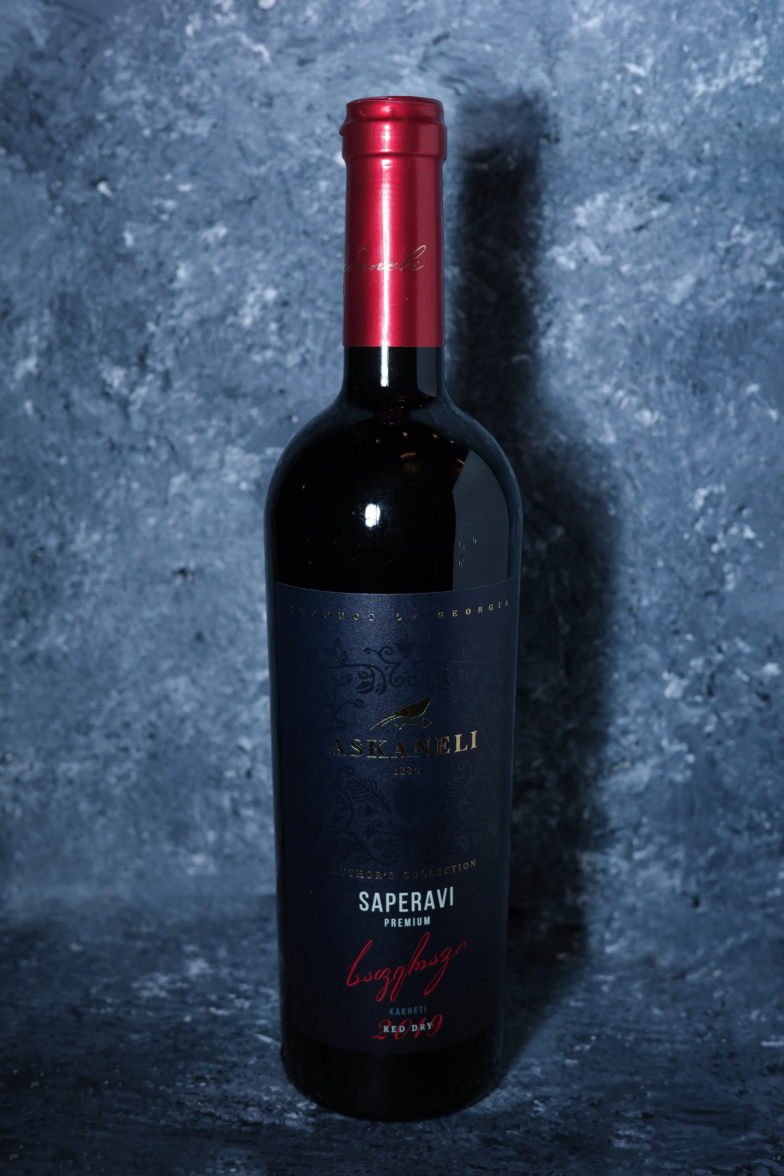 A dark-colored wine bottle with a red foil cap, label features gold and red text, set against a textured dark background.