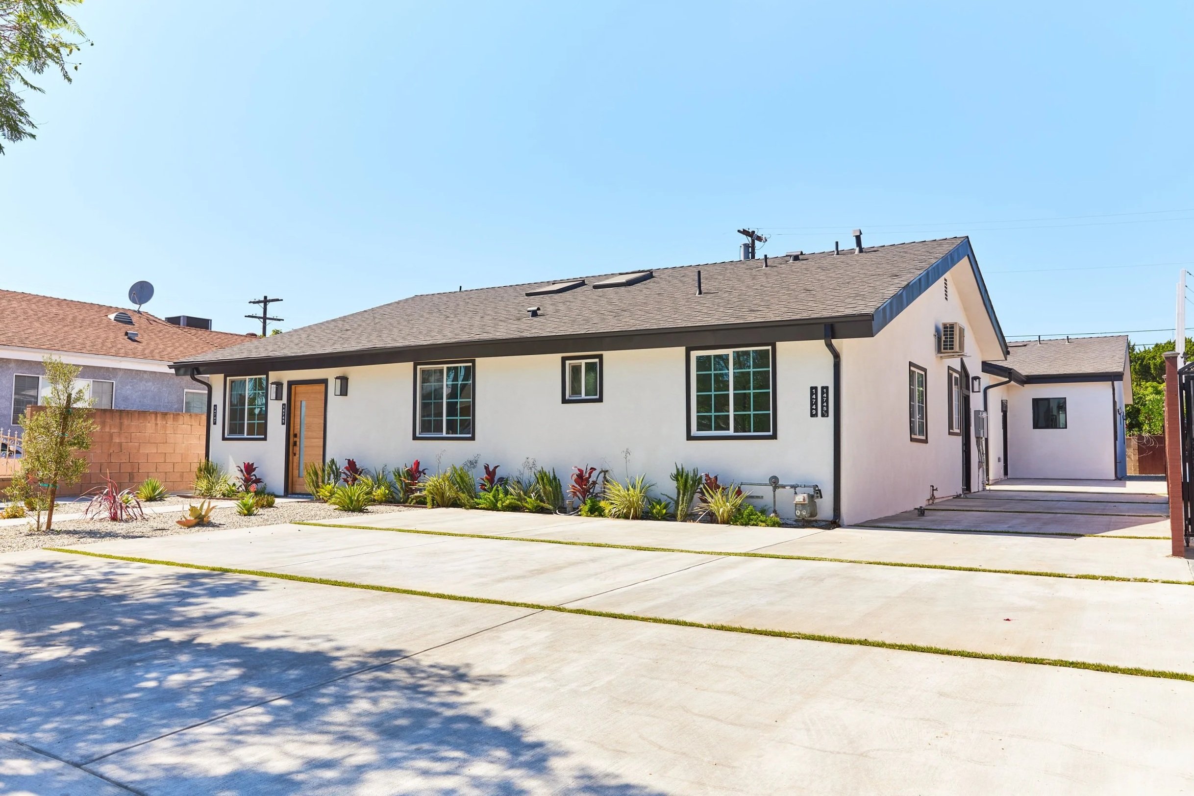 Exterior of a modern white house with black trim, brown front door, and landscaped front yard with plants and concrete driveway under a clear blue sky.