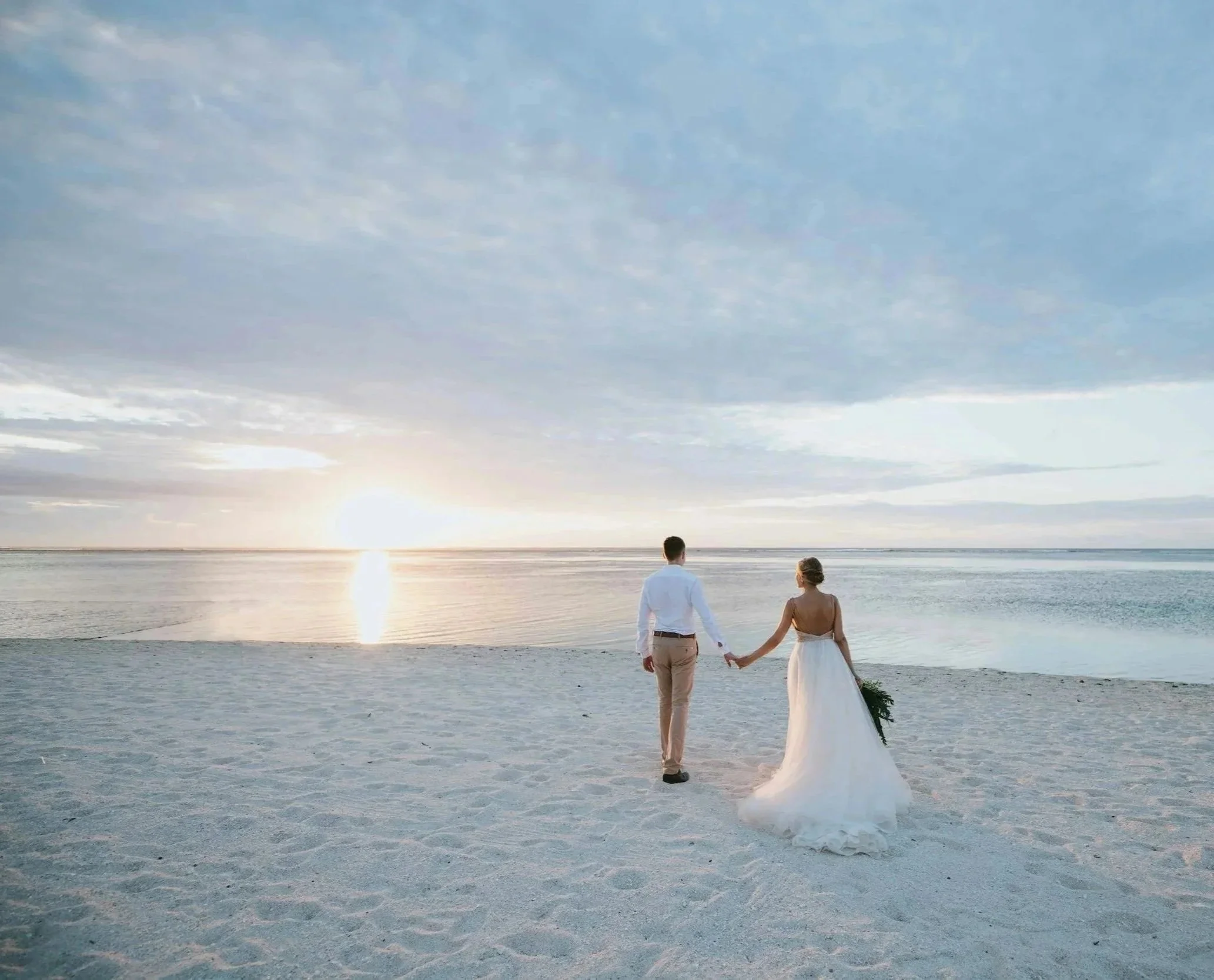 A bride and groom holding hands and walking on a sandy beach during sunset, with the ocean in the background.