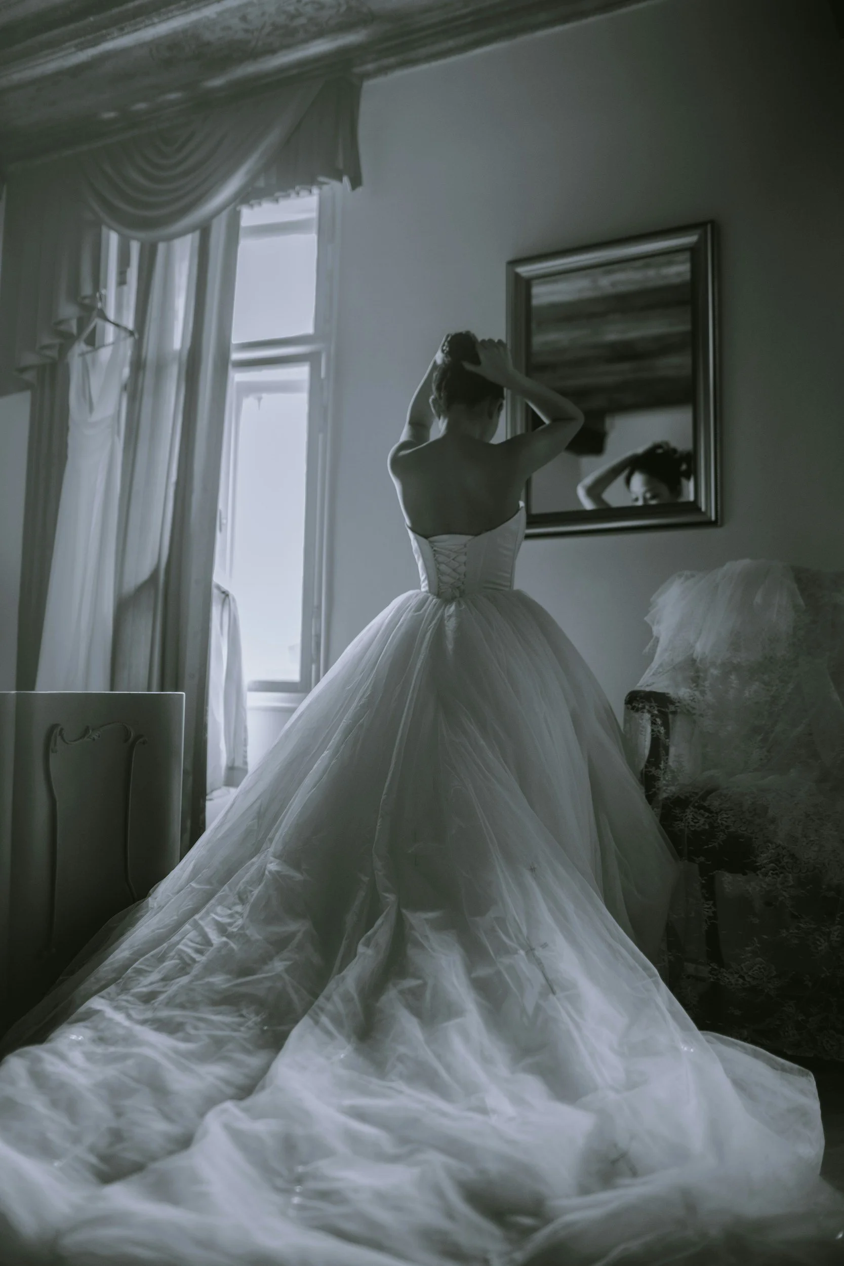 A woman in a strapless wedding gown with a full skirt is standing in a softly lit room, adjusting her hair in front of a mirror.
