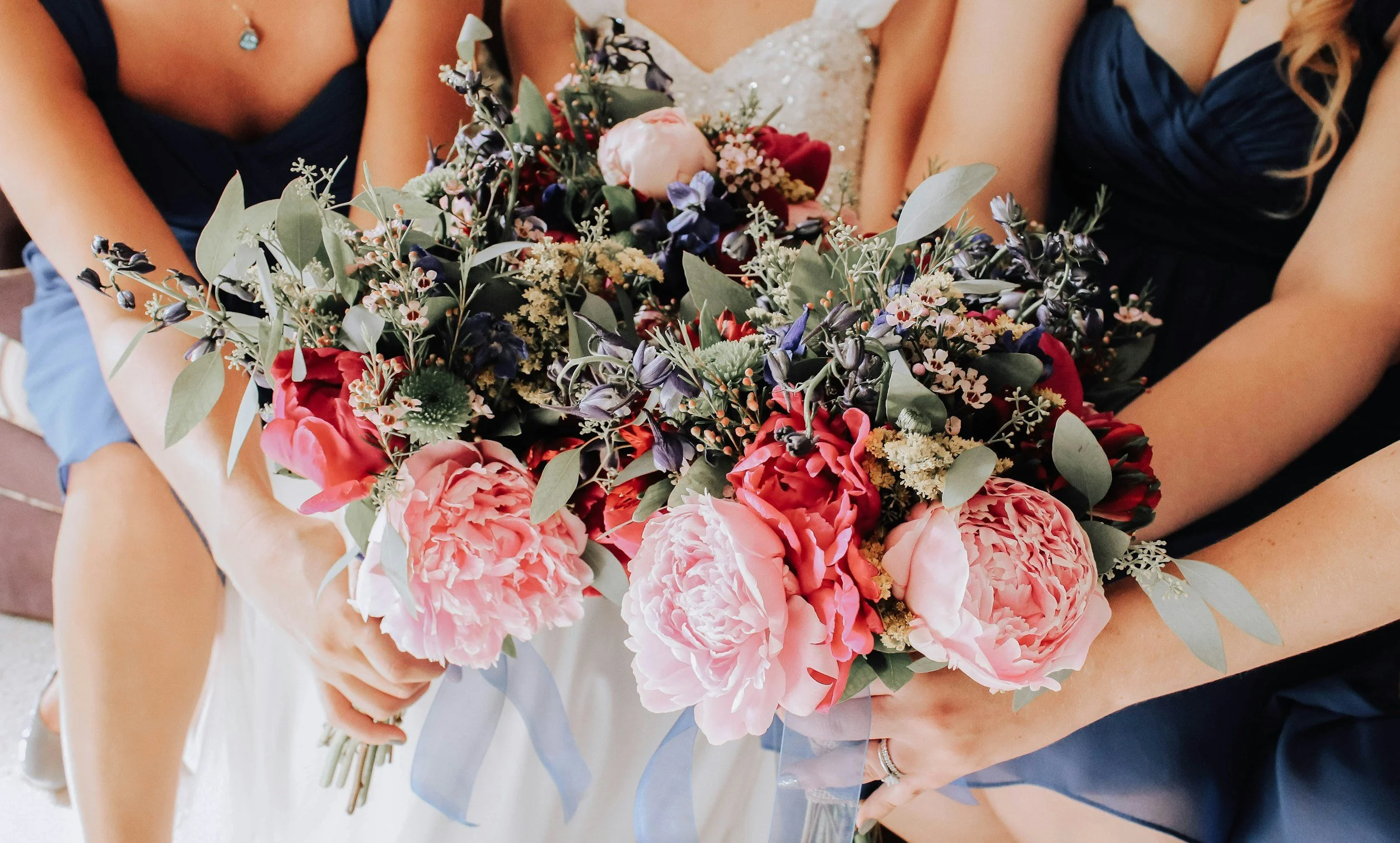 Close-up of a floral bouquet held by a bride and bridesmaids at a wedding. The bouquet includes pink peonies, red and purple flowers, and greenery.